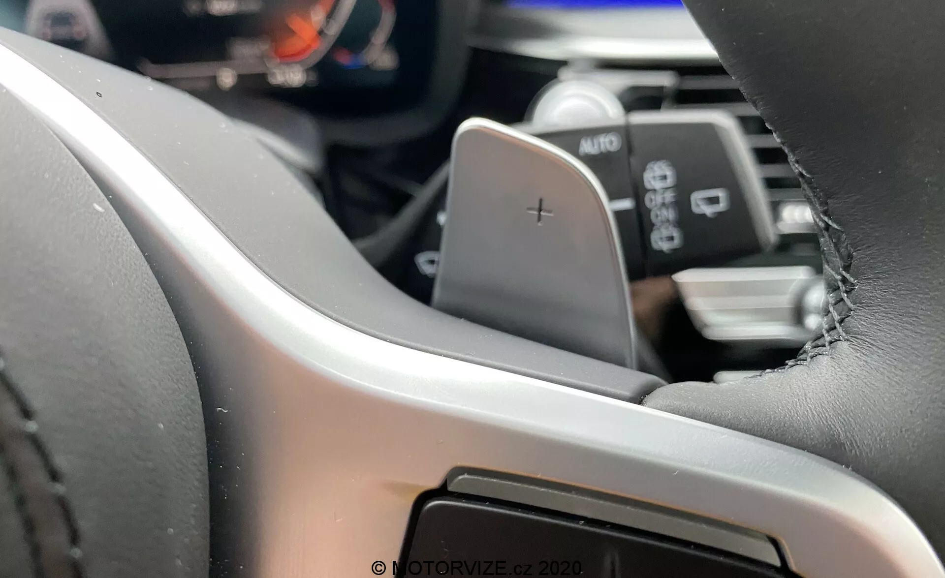 Interiérový pohľad na BMW 5 Series 2020 (BMW 520i Touring Steptronic) zameraný na ľavú stranu volantu z perspektívy vodiča⁠—moderný volant potiahnutý kožou s viditeľným šitím, vybavený páčkou na zmenu prevodov označenou '+'. Digitálny prístrojový panel alebo prístrojová doska je čiastočne viditeľná, čo zdôrazňuje pokročilé informačné systémy pre vodiča. Pod páčkou sa nachádza páčka s nápisom AUTO, ktorá pravdepodobne ovláda svetlá alebo stierače. V rozmazanom pozadí je viditeľná časť infotainmentového systému alebo centrálnej konzoly, čo naznačuje dotykové rozhranie.
