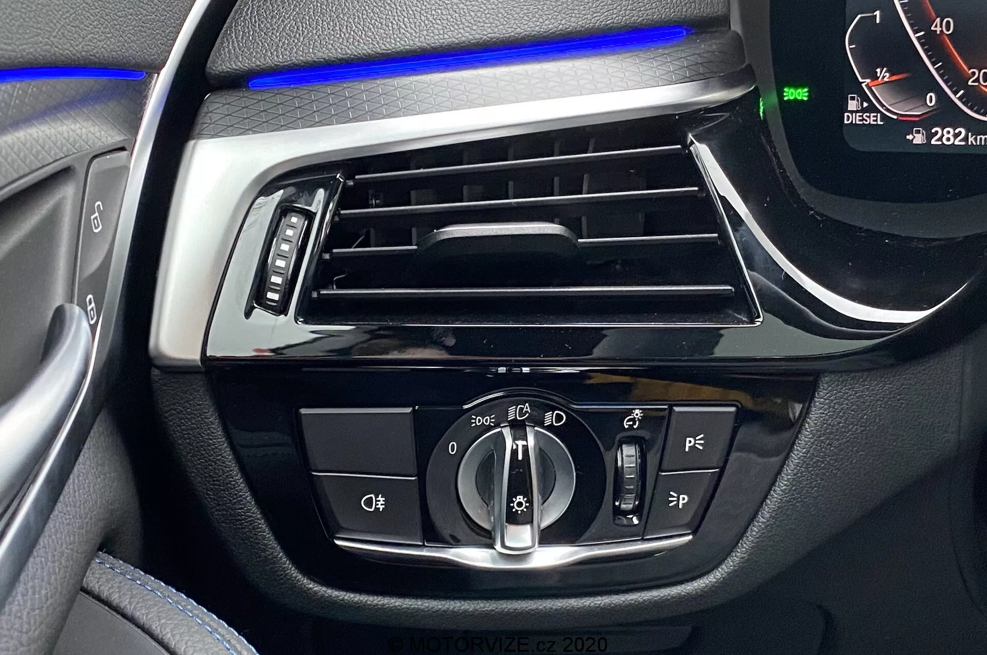 Záber interiéru BMW radu 5 2020, zameraný na prístrojovú dosku na strane vodiča s modrým ambientným osvetlením nad horizontálnym vetracím otvorom. Centrálne ovládanie zahŕňa otočný ovládač na nastavenie automatických svetiel a reflektorov, tlačidlá pravdepodobne pre hmlové svetlá a niveláciu reflektorov, ďalšie tlačidlá asistencie vodiča a digitálny tachometer ukazujúci 0 km/h s vozidlom v režime Park a nájazdom 282 km. Viditeľný je aj indikátor 'DIESEL'.