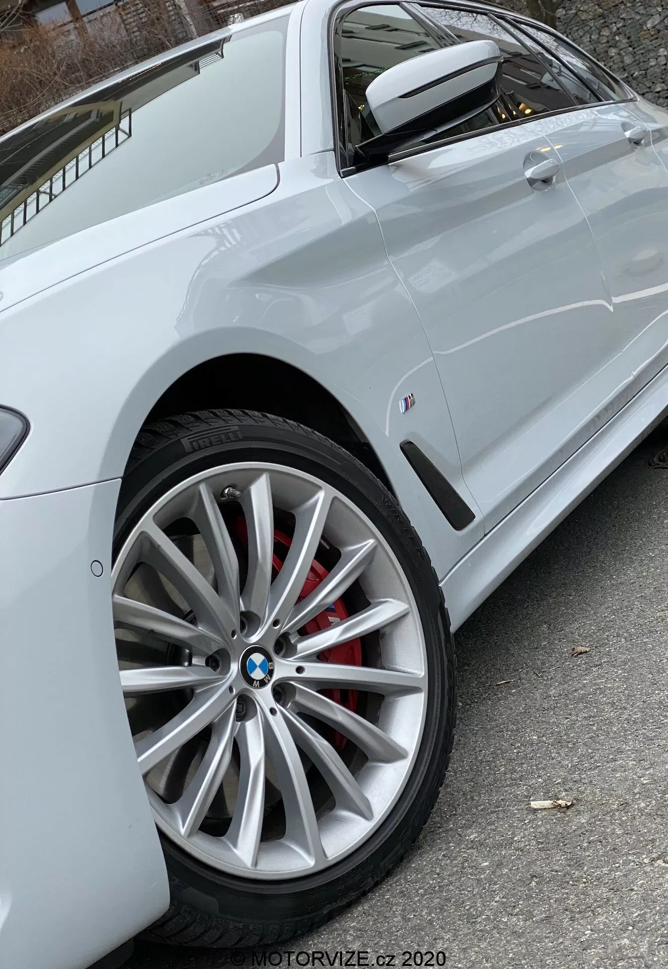 Detailný bočný pohľad na biely BMW 520i Touring Steptronic (2020) so zameraním na moderné viacramenné predné koleso s viditeľným logom BMW a červeným brzdovým strmeňom, čiastočne ukazujúci bočnú časť s ostrou charakterovou líniou cez kľučky dverí, farebne zladeným bočným zrkadlom s integrovaným smerovým svetlom a malým odznakom na prednom bočnom paneli. Prirodzené osvetlenie zaisťuje jasnú viditeľnosť vlastností vozidla. Foto od MOTORVIZE.cz 2020.