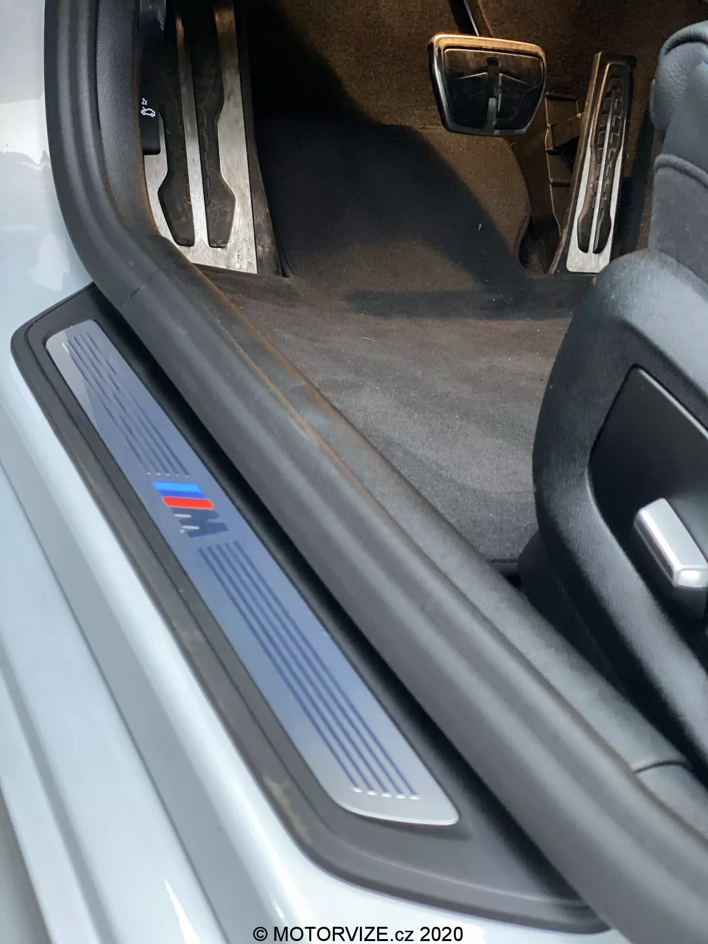 Zblúzka pohľad na prah dverí na vodičovej strane BMW 520i Touring Steptronic 2020. Obrázok ukazuje pohľad z nízkeho uhla, ktorý zvýrazňuje prahovú dosku z brúseného kovu s modrým prúžkom, čo môže naznačovať konkrétnu úroveň výbavy alebo športovú edíciu. Otvorené dvere nie sú viditeľné, ale ochranná a dekoratívna prahová doska je zrejmá, čo naznačuje možnú LED ilumináciu. Tmavý koberec interiéru a čiastočný pohľad na pedále s kovovým povrchom naznačujú športový interiérový balík. Na prahovej doske sú viditeľné zvyšky a znaky opotrebenia, čo naznačuje, že auto je používané.