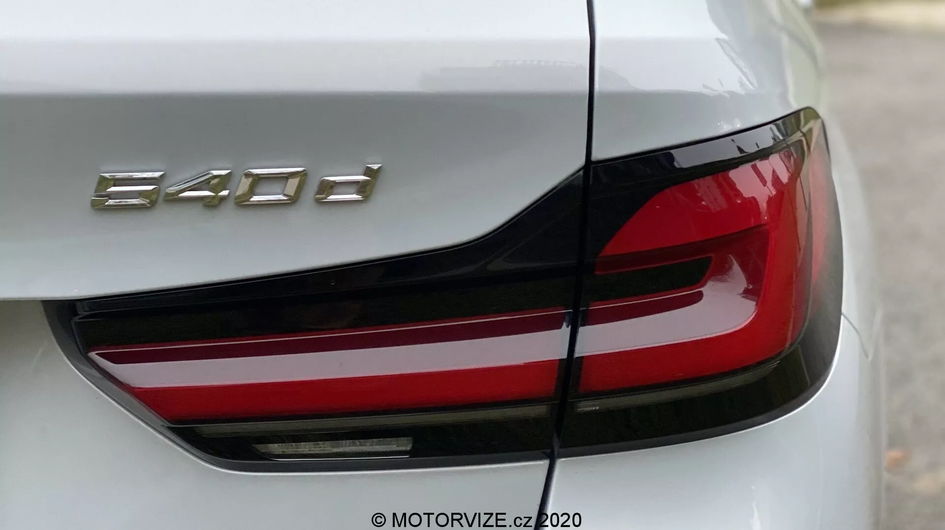 Detailný záber na zadnú časť BMW 520i Touring Steptronic so zameraním na ľavé zadné svetlo a časť kufra. Uhol je mierne šikmý z ľavého zadného rohu, zdôrazňujúci moderné LED zadné svetlo s ostrým, hranatým dizajnom a výraznou červenou časťou. Veko kufra ukazuje označenie modelu '840d.' Karoséria auta je lesklá, pravdepodobne biela alebo svetlo strieborná, zdôrazňujúca luxus a presné inžinierstvo.