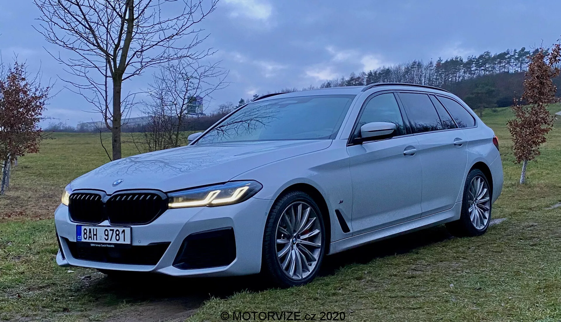 Obrázok luxusného kombi BMW radu 5 Touring 2020 (BMW 520i Touring Steptronic) v svetlosivej alebo šedej farbe, ktoré ukazuje agresívnu a športovú prednú časť s veľkými nasávacími otvormi, výraznou mriežkou a elegantnými LED svetlometmi. Vozidlo má veľké zliatinové kolesá s viaclúčovým dizajnom a výraznými brzdovými strmeňmi, dlhú strešnú líniu so spojlerom, chrómované okenné lišty a silnú bočnú líniu. Fotografia, ktorá bola zhotovená vo výške stredu vozidla v polovenkovskom prostredí s difúznym osvetlením.