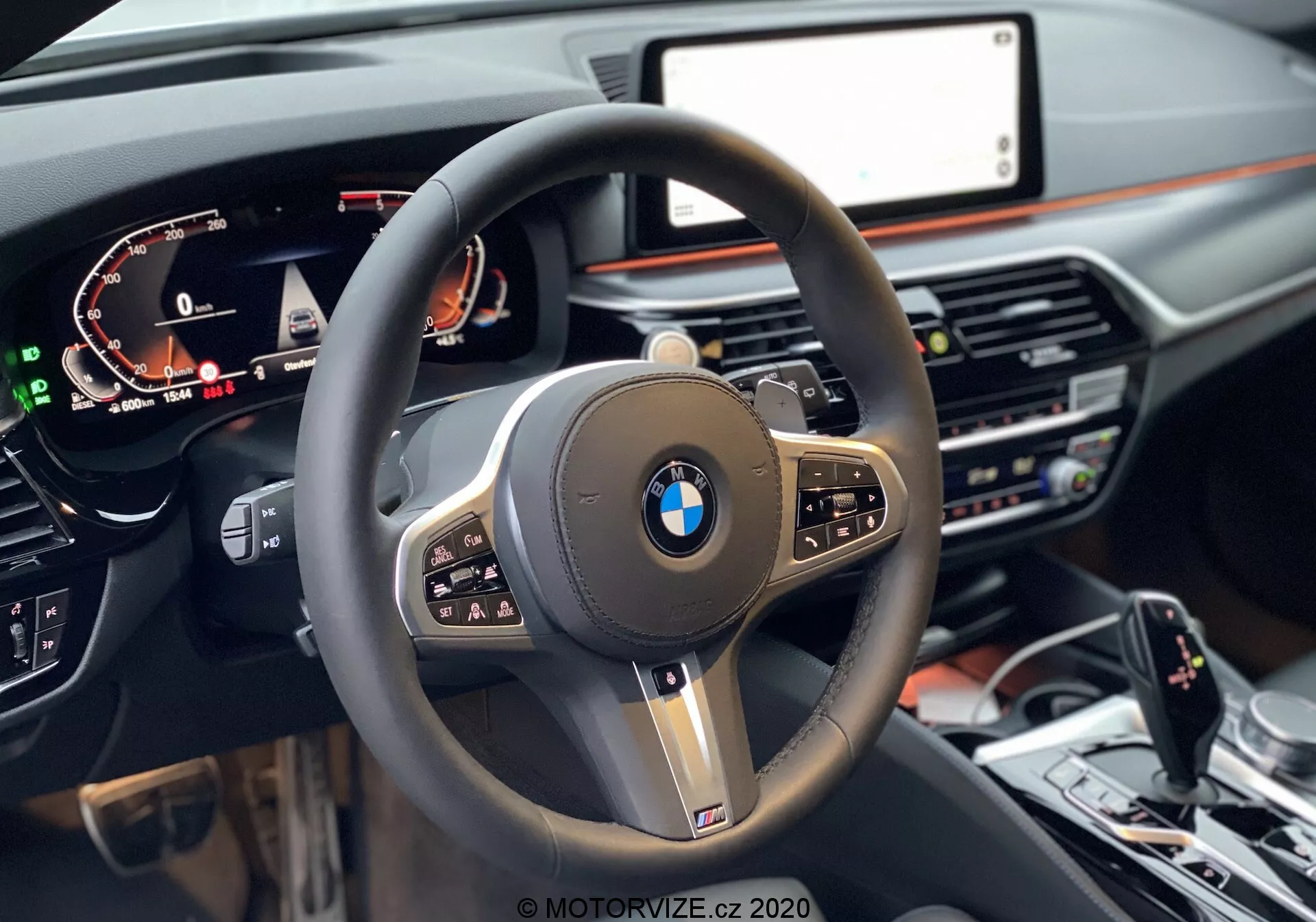 Pohľad z prvej osoby zo sedadla vodiča v BMW 520i Touring Steptronic 2020, zameraný na moderný trojramenný volant s hrubým koženým obalom, multifunkčnými tlačidlami a výrazným logom BMW v strede, obklopeným lesklými čiernymi a kovovými akcentmi. Digitálny prístrojový panel na ľavej strane zobrazuje rýchlosť, otáčky motora a informácie pre vodiča, zatiaľ čo na pravej strane je čiastočne viditeľná centrálna informačná obrazovka. Palubná doska tmavých tónov s pridaním dreva obsahuje pod obrazovkou ovládanie klimatizácie a systém médií. V spodnej časti obrázka vidíme stredovú konzolu s voličom rýchlostí, iDrive ovládačom a tlačidlami na ovládanie dynamiky jazdy.