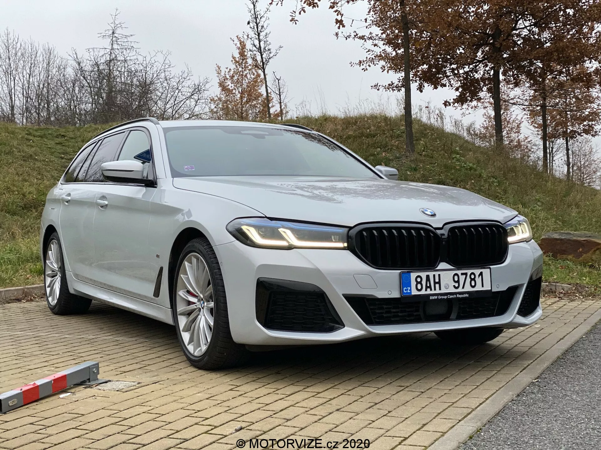 Obrázok zobrazuje BMW 5er-reihe 2020 (520i Touring Steptronic), moderný luxusný sedan, zaparkovaný na miernom svahu. Snímka je urobená z nízkeho a mierne šikmého uhla spredu, čo zvýrazňuje prednú štvrtinu a profil vozidla. Výrazné detaily zahŕňajú prominentnú mriežku v tvare obličky, komplikované prívody vzduchu na prednom nárazníku, elegantné, uhlové LED svetlomety, veľké viacramenné zliatinové kolesá, dlhú, plynulú siluetu karosérie s kovovo striebornou úpravou, chrómové lišty, integrované smerovky na bočných zrkadlách a potenciálne pokročilé technológie ako autonómne jazdné asistenty a asistenčný parkovací systém. Na prednom štvrtinovom paneli je viditeľný odznak, ktorý označuje konkrétnu variantu modelu.