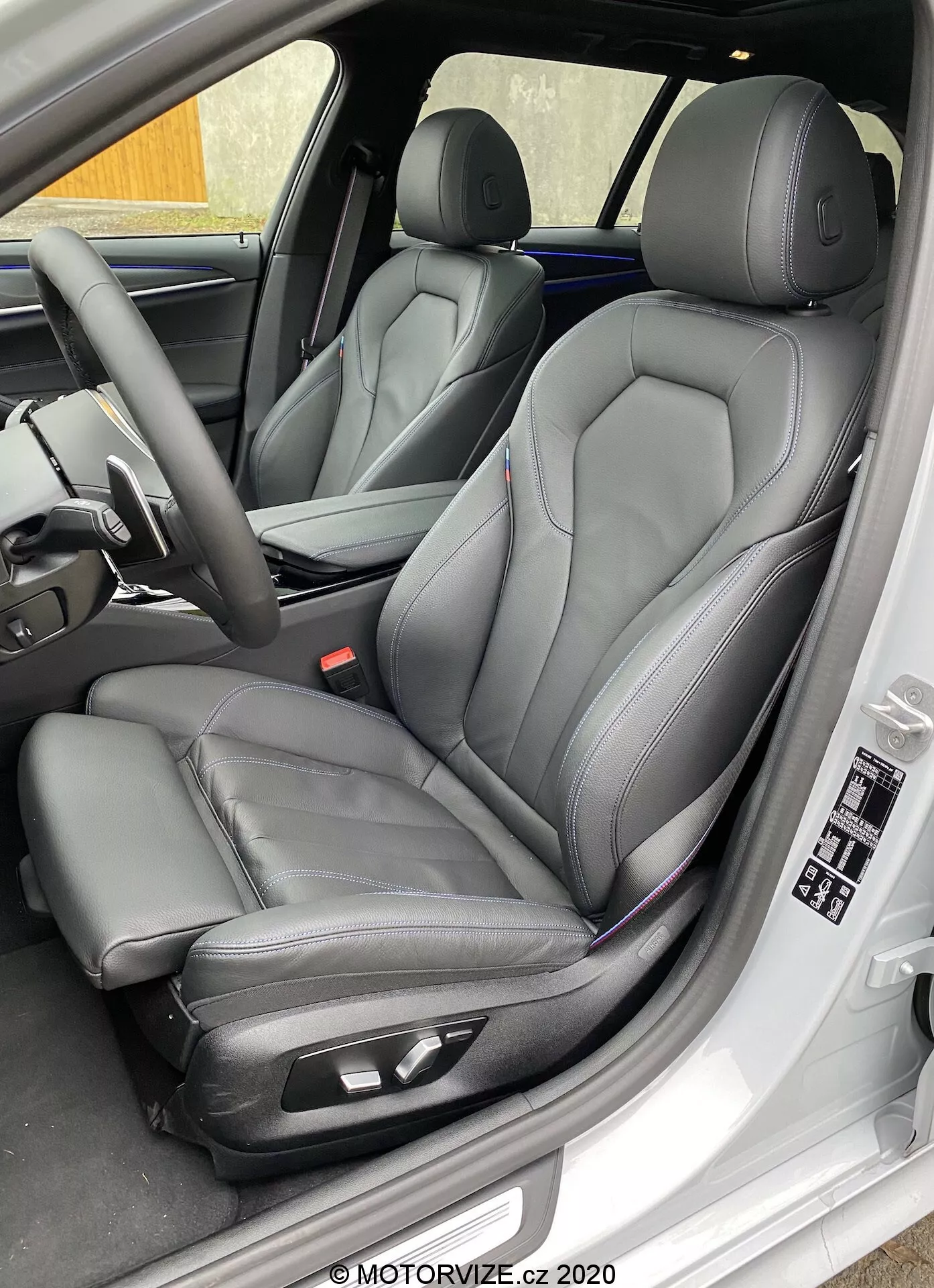 Detailný záber z bočného uhla na sedadlo vodiča modelu BMW 520i Touring Steptronic 2020, ukazujúci moderné predné sedadlá štýlu vedra v čiernej koži s kontrastným prešívaním, vybavené elektronickými ovládacími prvkami pre zlepšený komfort. Obrázok zachytáva aj vnútorný panel dverí vodiča s elektrickými ovládačmi okien a zrkadiel, nastavenia pamäte sedadla a ďalšie prvky prístrojovej dosky a volantu, ktoré naznačujú luxus a pokročilú technológiu.