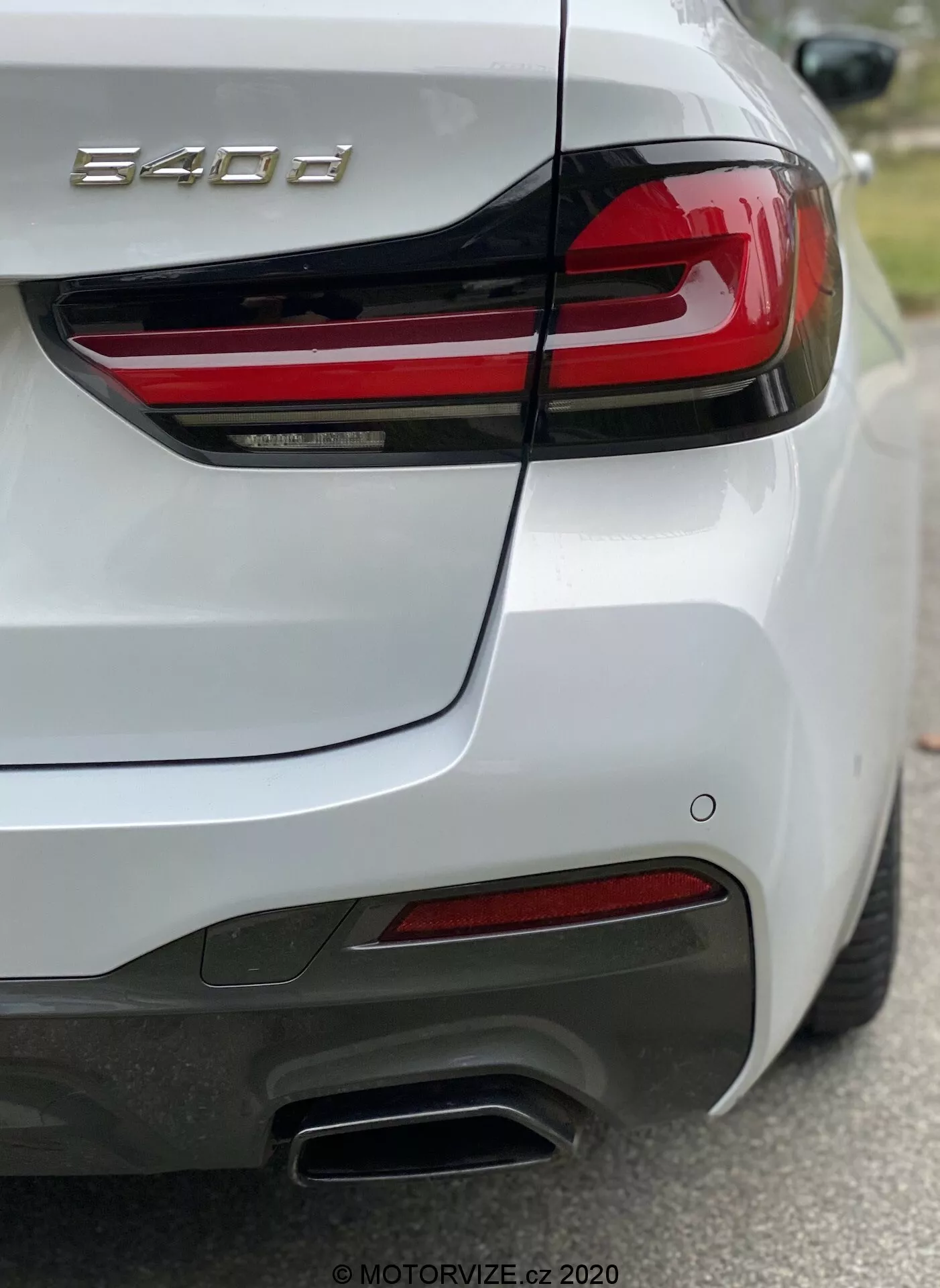 Zadná časť osobného vozidla BMW radu 8 (840i), zachytená zo zadného pravého uhla, zvýrazňujúc pravé zadné svetlo s vodorovnými červenými LED pruhmi, tvarované veko kufra s integrovaným spojlerom, zadný nárazník so senzormi, dvojité kovové výfuky na pravej strane, červené odrazky a lesklý biely lak. Fotografia sa zameriava na znak '840i', ukazuje detailné prvky dizajnu a technológie, ako sú osvetlenie a asistenčné systémy vodiča.
