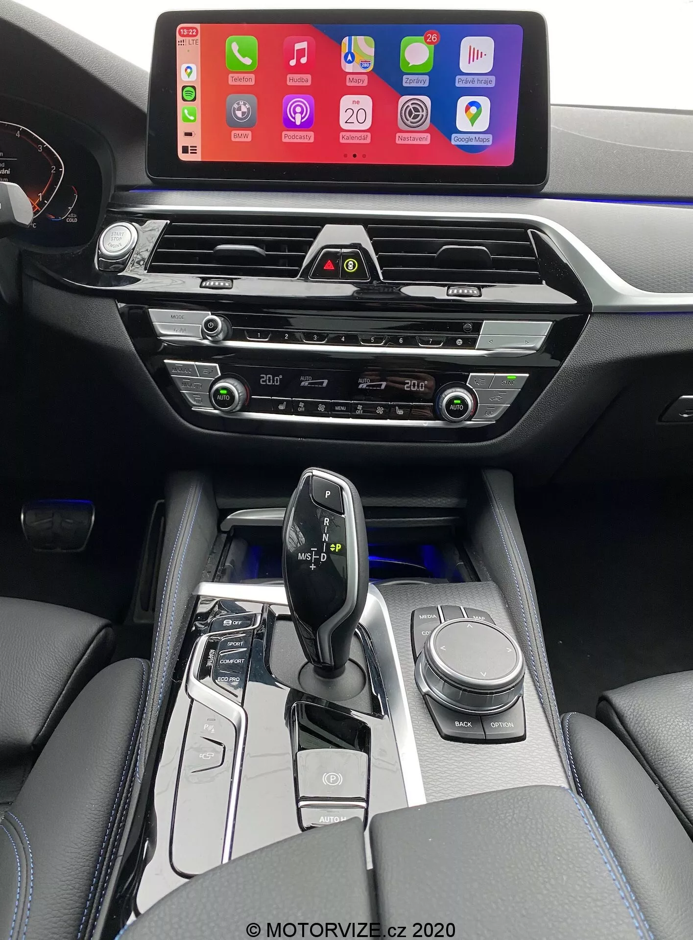 Interiérový pohľad na BMW 520i Touring 2020 s prevodovkou Steptronic, zo sedadla vodiča mierne vyvýšený, aby bol jasnejší pohľad na stredovú konzolu, oblasť radiacej páky a časť palubnej dosky. Obrázok zobrazuje veľkú infotainment obrazovku zobrazujúcu rozhranie pre pripojenie smartfónu, pravdepodobne Apple CarPlay, namontovanú vysoko na palubnej doske pre ľahkú viditeľnosť. Pod obrazovkou sú moderné horizontálne ventilačné otvory s chrómovými akcentmi, následované hmatovými tlačidlami a otočnými ovládačmi na ovládanie klímy. Stredová konzola obsahuje aj ovládanie audio systému, ďalšie ovládače klímy a možný slot pre CD prehrávač. Ergonomická a minimalistická radiaca páka, pravdepodobne elektronická, je obklopená vysokolesklým čiernym a kovovým lemovím, s tlačidlami na výber jazdného režimu a otočným ovládačom na navigáciu. Interiér tiež obsahuje čalúnené opierky s viditeľným prešívaním, čo pridáva na luxusnom pocite. Mäkké biele osvetlenie zvýrazňuje tlačidlá okolo oblasti radiacej páky, čo naznačuje automatickú prevodovku.