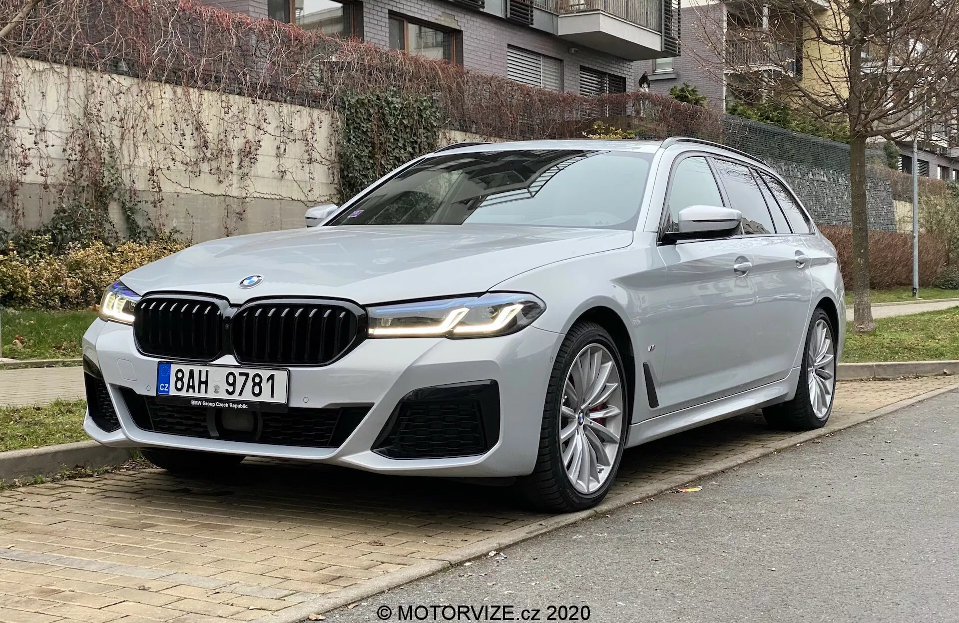Luxusné BMW 520i Touring zaparkované na mestskej ulici, pohľad z nízkeho čelného trojštvrťového uhla zvýrazňuje jeho prednú časť a stranu vodiča. Auto má výraznú mriežku s chromatickými detailmi, štýlové LED svetlomety, športový predný nárazník a výrazné záhyby kapoty. Viaclúčové hliníkové kolesá, bočné zrkadlá s ukazovateľmi smeru a kamerou, chrómové rámy okien a strešné okno pridávajú luxusný vzhľad. Umiestnené na pravej strane s trávou, kríkmi a obytnými budovami v pozadí, naznačujúc pokročilú technológiu a špičkový dizajn.