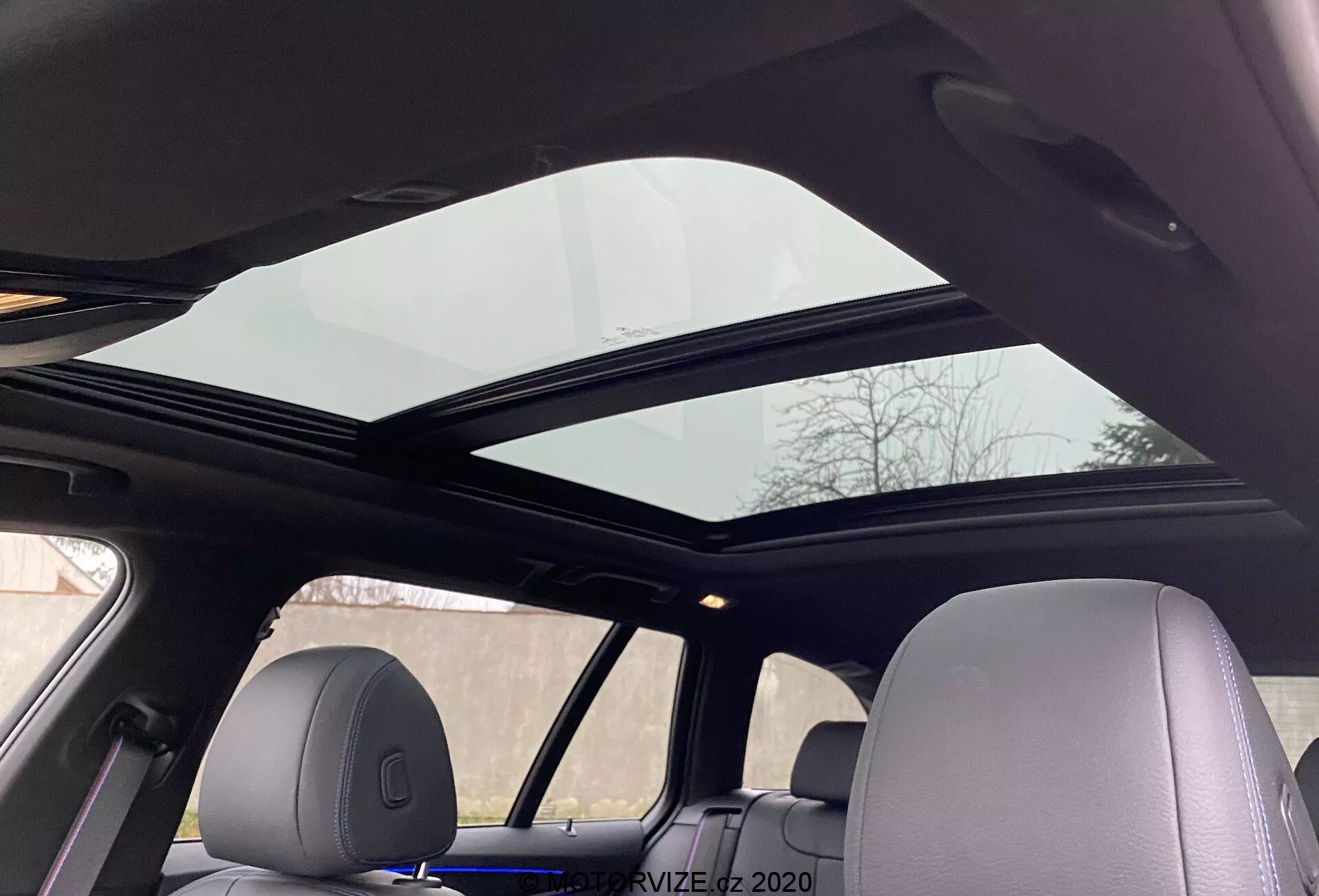 Interiér BMW radu 5 2020 520i Touring so Steptronic prevodovkou, ktorá ukazuje panoramatické strešné okno rozprestreté nad prednými a zadnými sedadlami z pohľadu sediaceho na zadnom sedadle smerom nahor. Predný panel strešného okna je mierne otvorený, čo zlepšuje vetranie. Stropné obloženie je svetlé, dopĺňajúce konzistentný dizajn interiéru, s opierkami hlavy zodpovedajúcimi materiálu interiéru, zdôrazňujúcimi pokročilý komfort a moderné vybavenie.