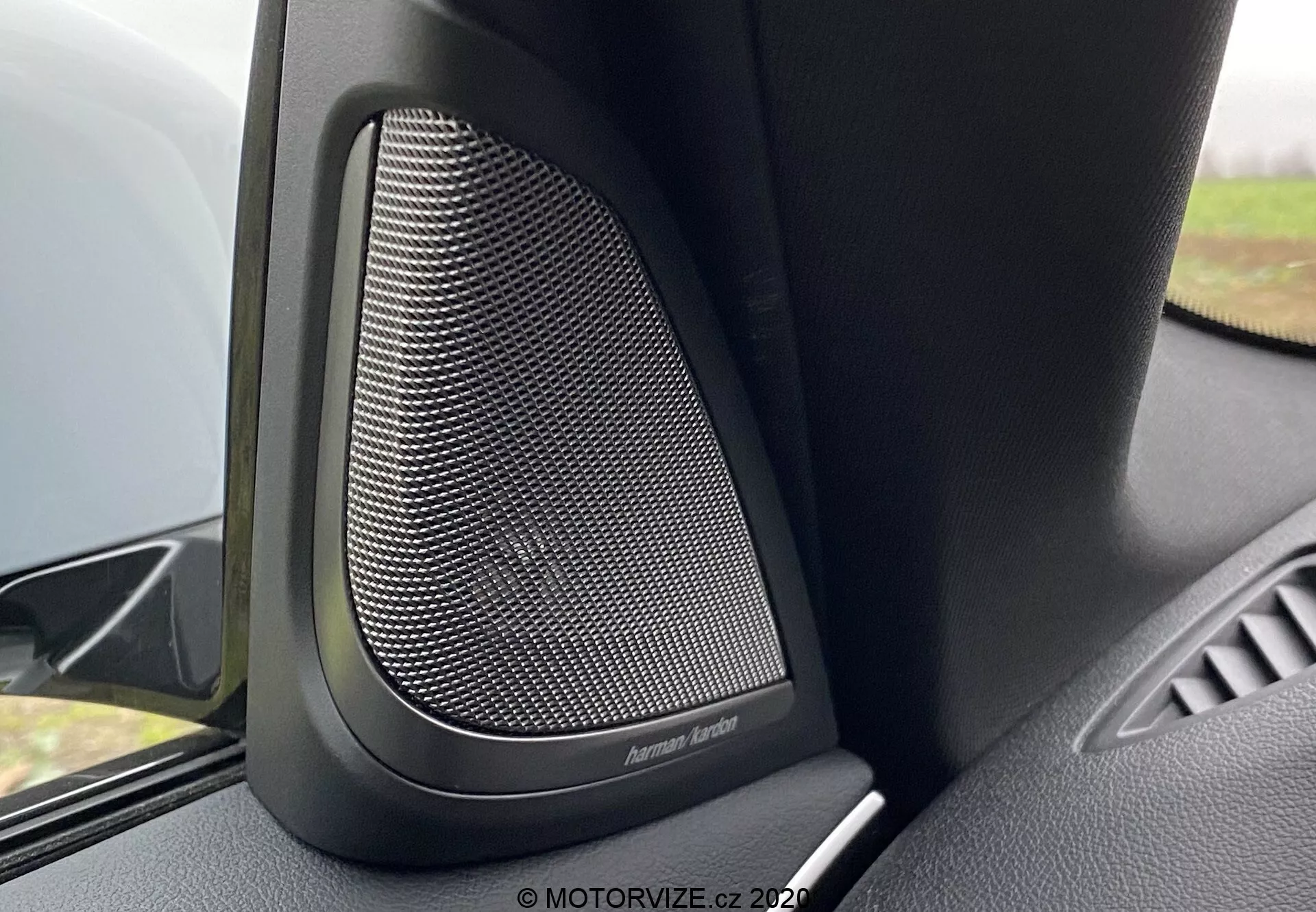 Zbližší pohľad na interiér BMW 5er-Reihe 2020 (BMW 520i Touring Steptronic) so zameraním na prémiový dverový reproduktor Harman Kardon. Obrázok zachytáva jemný sieťový vzor kovovej mriežky reproduktora, obklopenej čiernym plastom alebo mäkkými materiálmi určenými na tlmenie vibrácií pre vynikajúcu kvalitu zvuku. Uhol kamery ukazuje hornú časť dverného panelu s výrazným reproduktorom v popredí a časťou opierky dverí. Mierne rozostrenie pozadia zdôrazňuje túto high-end audio vlastnosť v tomto luxusnom vozidle.