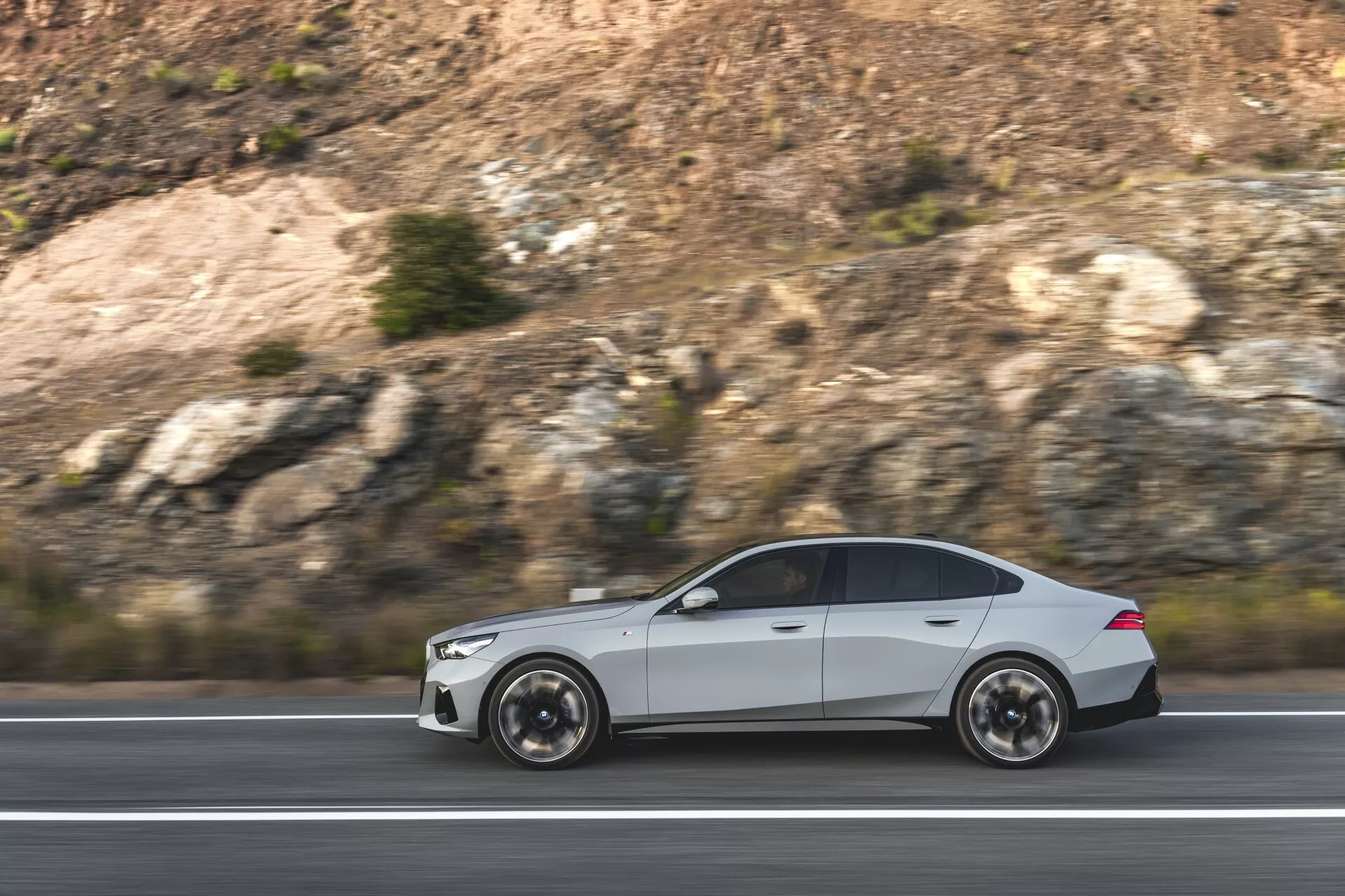 BMW radu 5 (G60-G61) 2023 v pohybe, ukazujúce jeho aerodynamický dizajn a športovú estetiku s viditeľnými viacramennými kolesami a elegantnými kontúrami karosérie, zachytené z boku na diaľnici