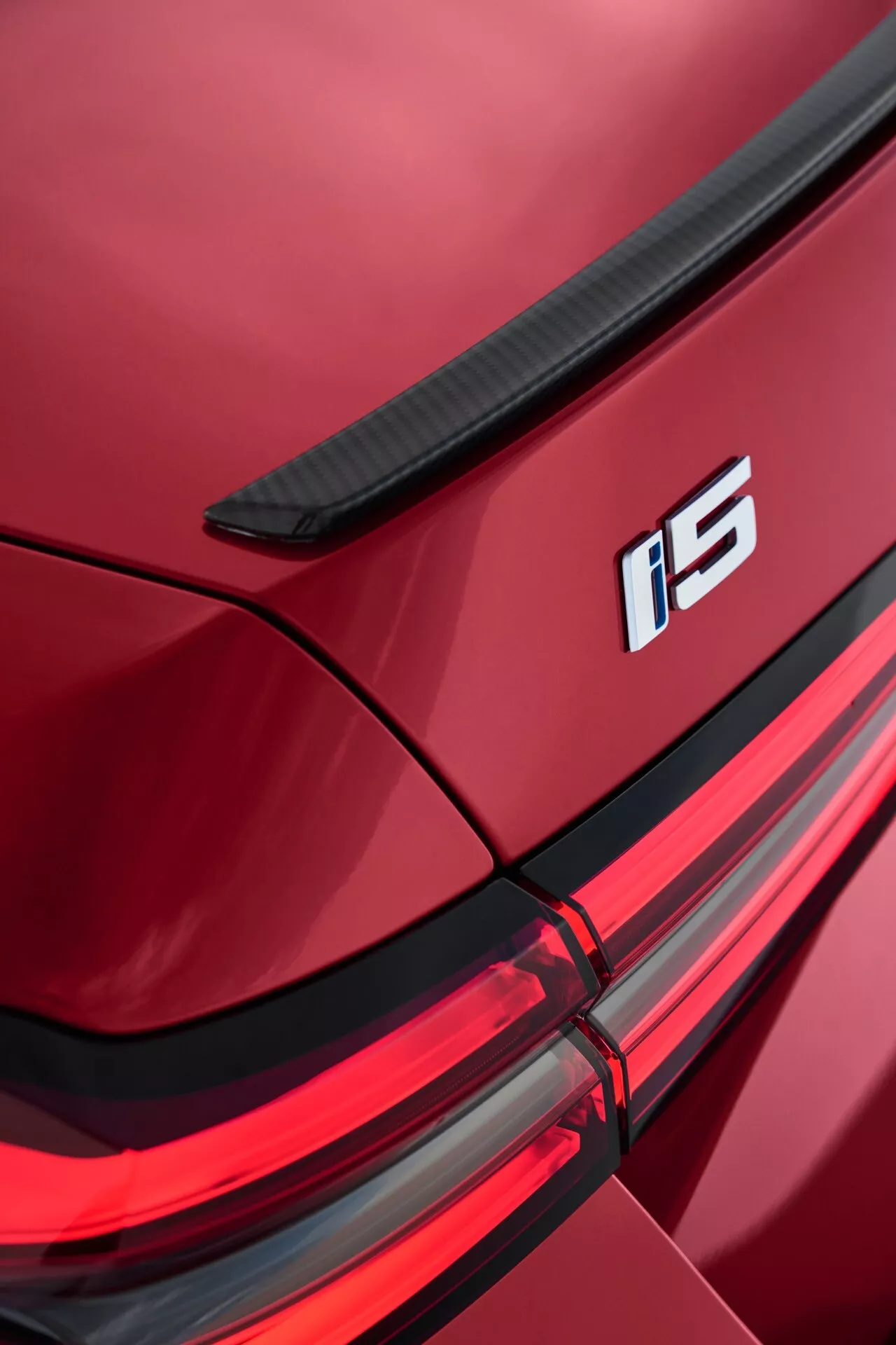 Detailný pohľad na pravý zadný roh BMW rady 5 (G60-G61) 2023 s integrovaným spoilerm na veku batožinového priestoru, označením i5, súpravou LED zadných svetiel a lesklým metalickým červeným lakováním