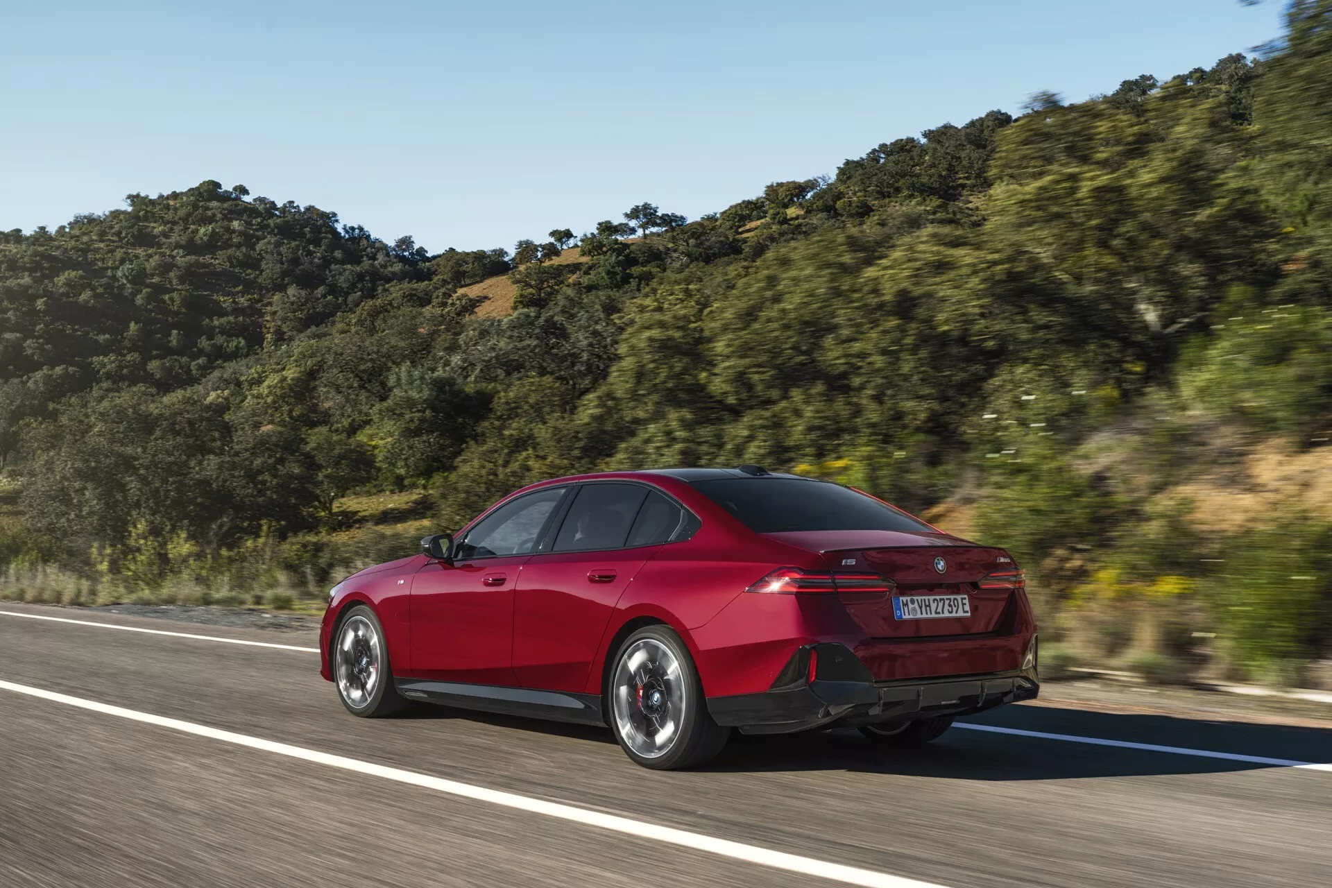 Červený sedan BMW radu 5 (G60-G61) so štyrmi dverami v pohybe zachytený z trojštvrťového zadného uhla, zdôrazňujúci zadné a bočné dizajnové prvky ako tvar kufra, zadný nárazník, LED zadné svetlá a aerodynamické línie na pozadí horskej krajiny pod jasnou oblohou