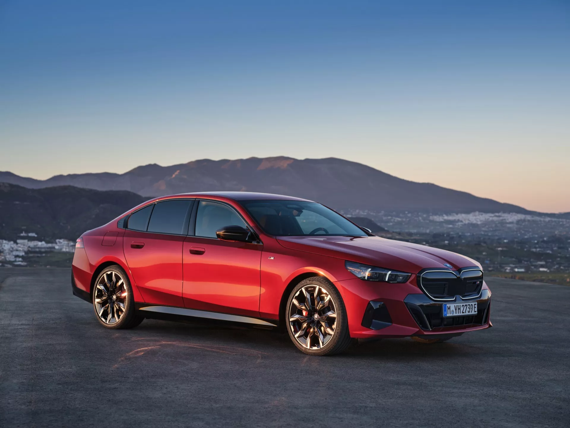 BMW radu 5 z roku 2023 (G60-G61) zobrazené pod uhlom tri štvrtiny zpredu, zdôrazňujúce jeho charakteristický predný gril s logom uprostred, úzke LED svetlomety, predný nárazník s integrovanými vzduchovými otvormi a leštené kolesá z ľahkých zliatin s viacerými lúčmi proti dynamickému červenému metalickému finišu a pozadiu súmraku