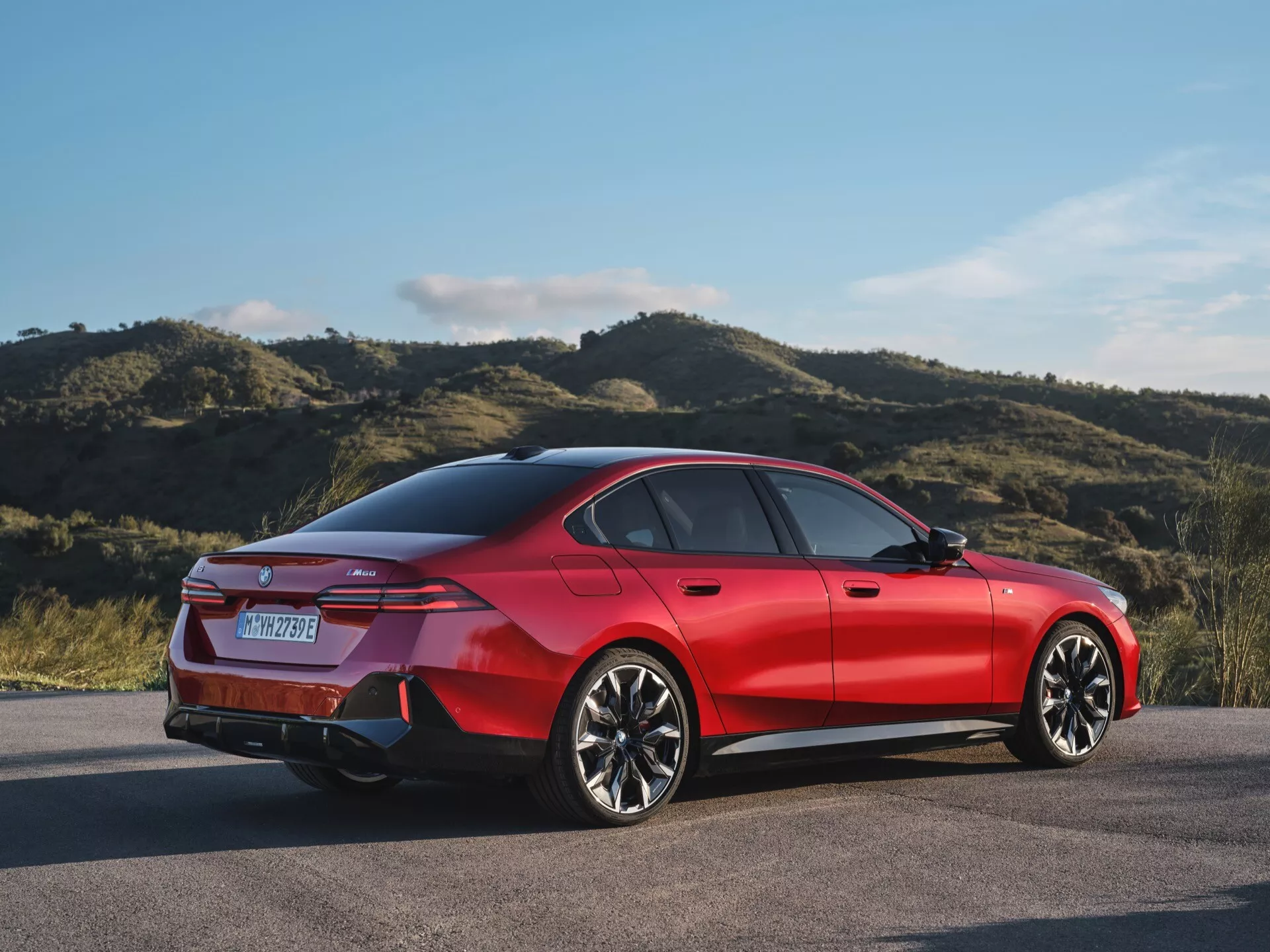 Červený štvordverový sedan BMW radu 5 (G60-G61) z roku 2023, zaparkovaný vonku, ktorý ukazuje jeho zadný trojštvrťový profil, zdôrazňujúci športový postoj vozidla, moderný podpis LED zadných svetiel, disky z ľahkej zliatiny s viacerými ramenami a siluetu pripomínajúcu kupé.