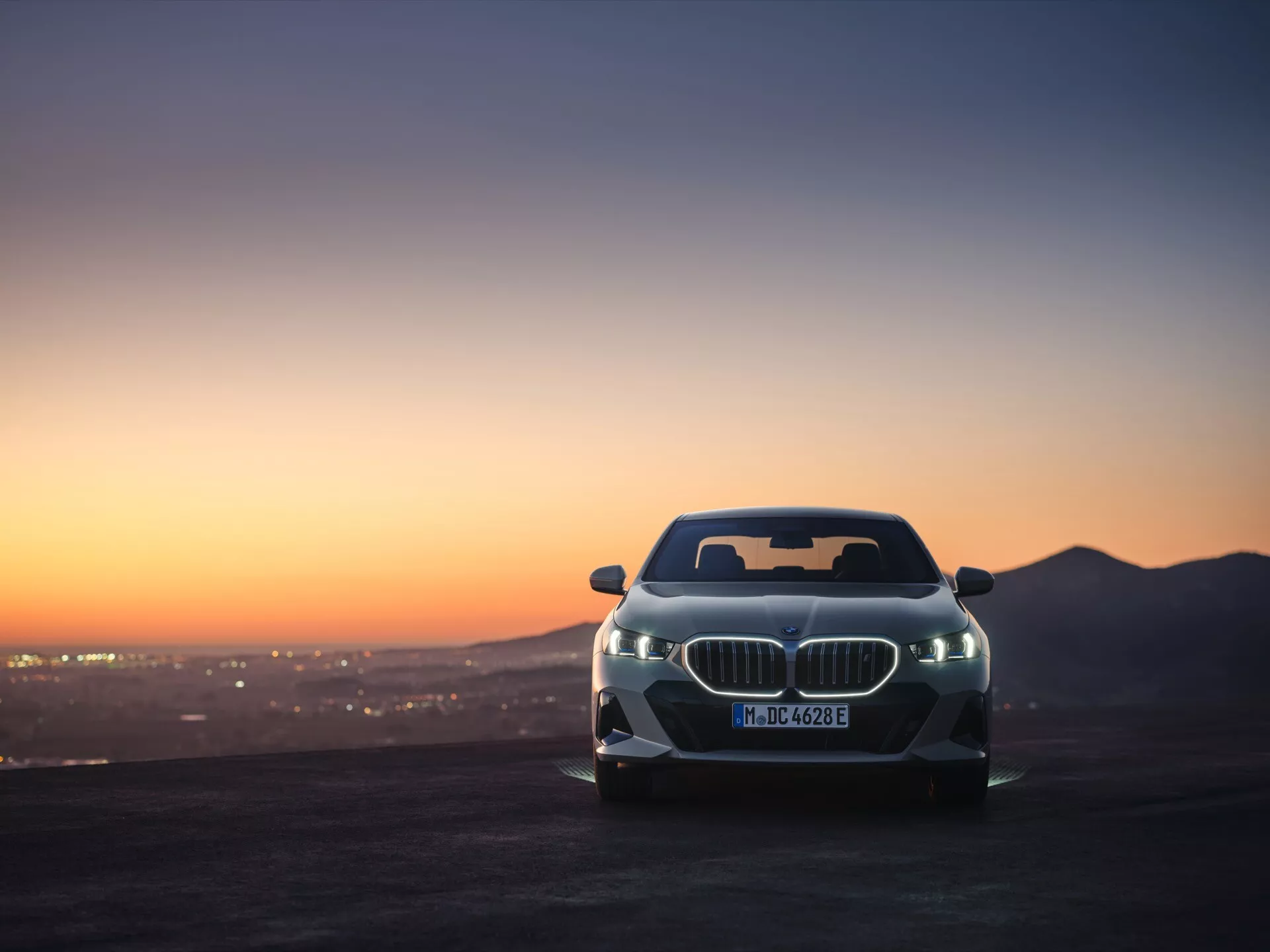 Uhlový predný pohľad na model BMW série 5 (G60-G61) 2023 s ikonickou obličkovou mriežkou a pokročilými LED svetlami počas malebnej svitania alebo západu slnka, predvádzajúci aerodynamický dizajn vozidla a luxusnú estetiku na pozadí hornatého terénu a žiariacej mestské krajiny.