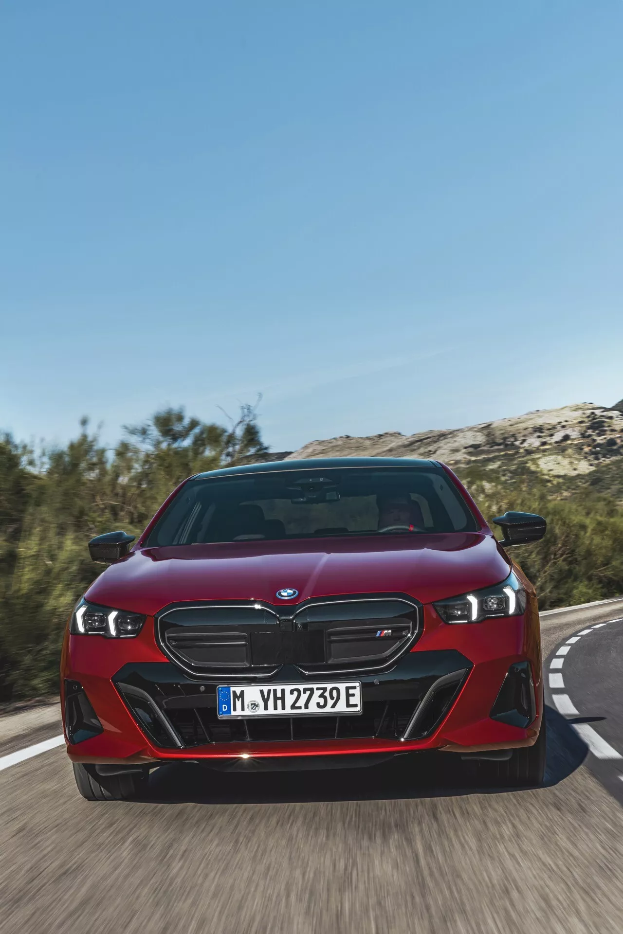 BMW radu 5 (G60-G61) 2023 v červenej farbe, predvádzajúce charakteristickú obličkovú mriežku, LED svetlomety a dynamický predný nárazník z nízkeho uhla spredu na ceste, čo zdôrazňuje rýchlosť vozidla a pokročilé elektrifikované vlastnosti iPerformance