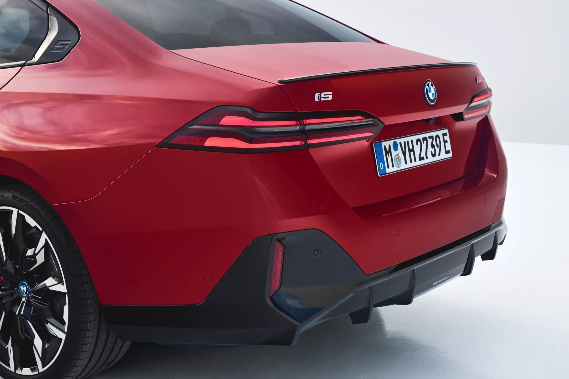 Zadný uhlový pohľad na červené BMW i5 2023 zobrazujúce športový sedanový dizajn, integrovaný spoiler, výrazný znak BMW, predĺžené LED zadné svetlá siahajúce do zadných blatníkov, označenie elektrického modelu 'i5', agresívny zadný nárazník s dekoratívnym difúzorom a falošnými výfukmi, v trojštvrťovom zadnom a bočnom profile.