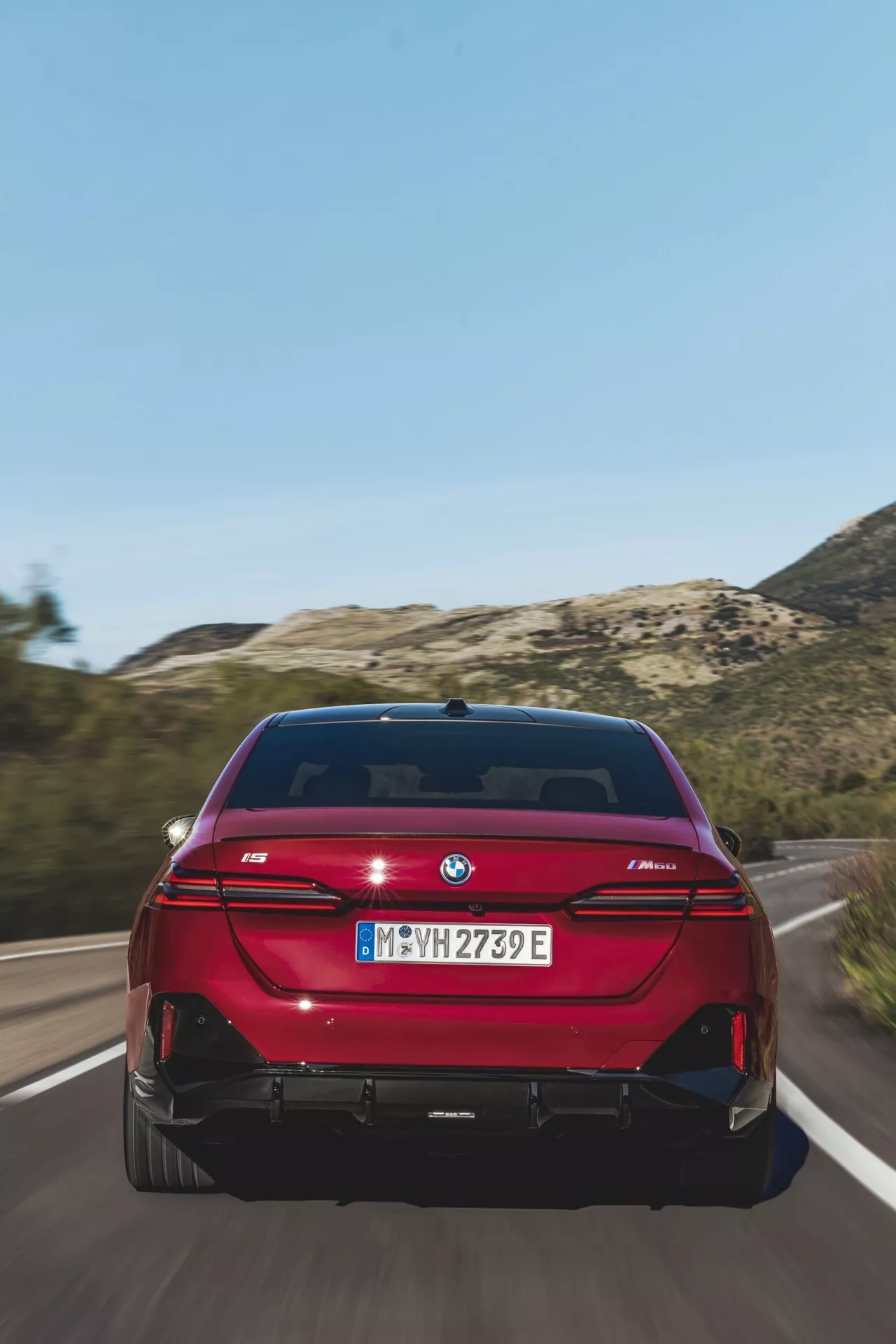 Zadný pohľad na žiarivo červený sedan BMW radu 5 (G60-G61) idúci po ceste, s LED zadnými svetlami v tvare L, duálnym výfukovým systémom, odznakom eDrive a parkovacími senzormi pri jasnom dennom svetle.