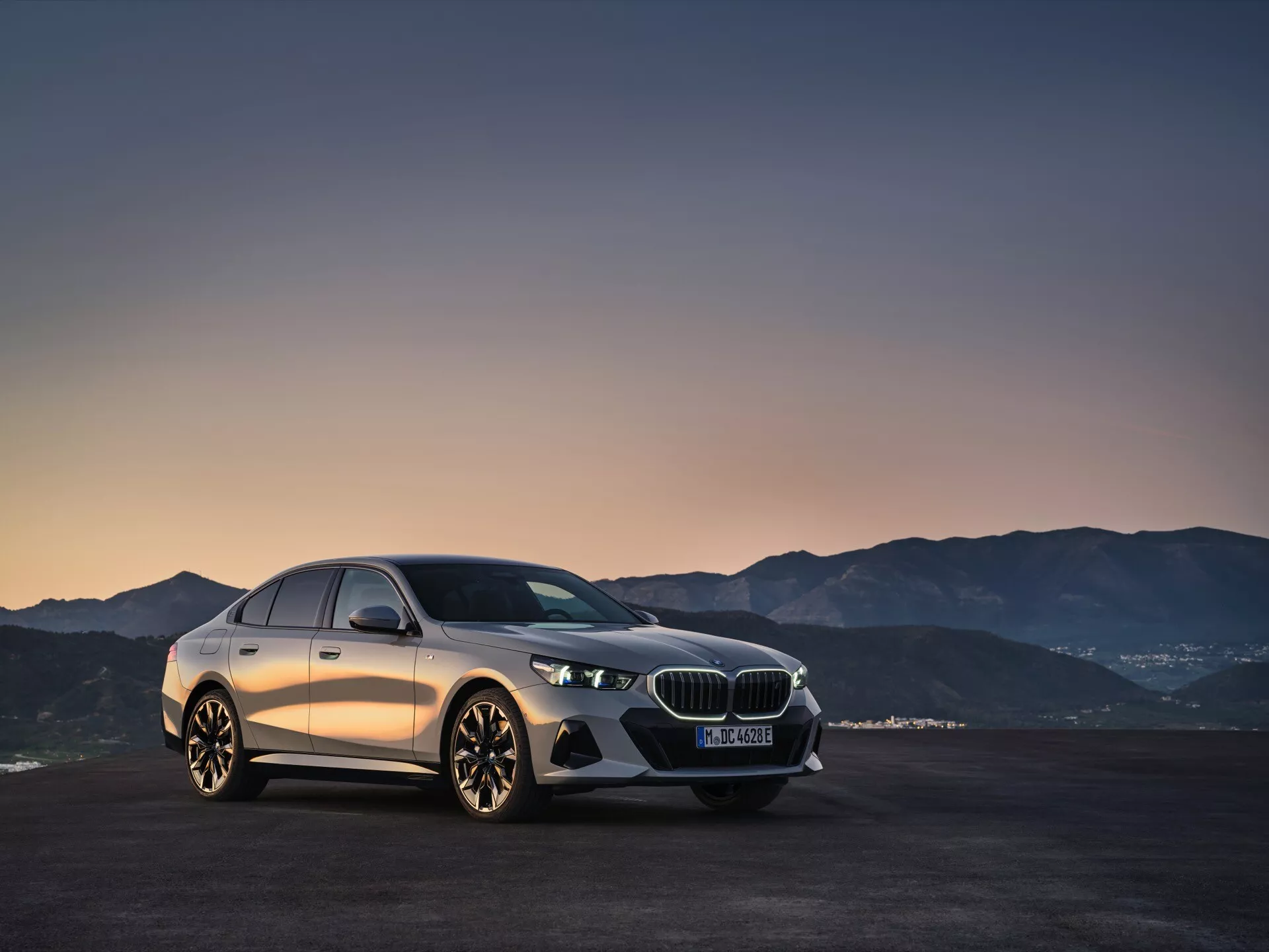 BMW radu 5 (G60-G61) 2023 predvádzané na úsvite alebo súmraku, zobrazujúce úplný pohľad na jeho aerodynamický dizajn, zdôraznený pokročilým systémom LED svetlometov, luxusnou prednou mriežkou so znakom a detailným bočným profilom na pozadí gradientu teplého neba a horskej krajiny.