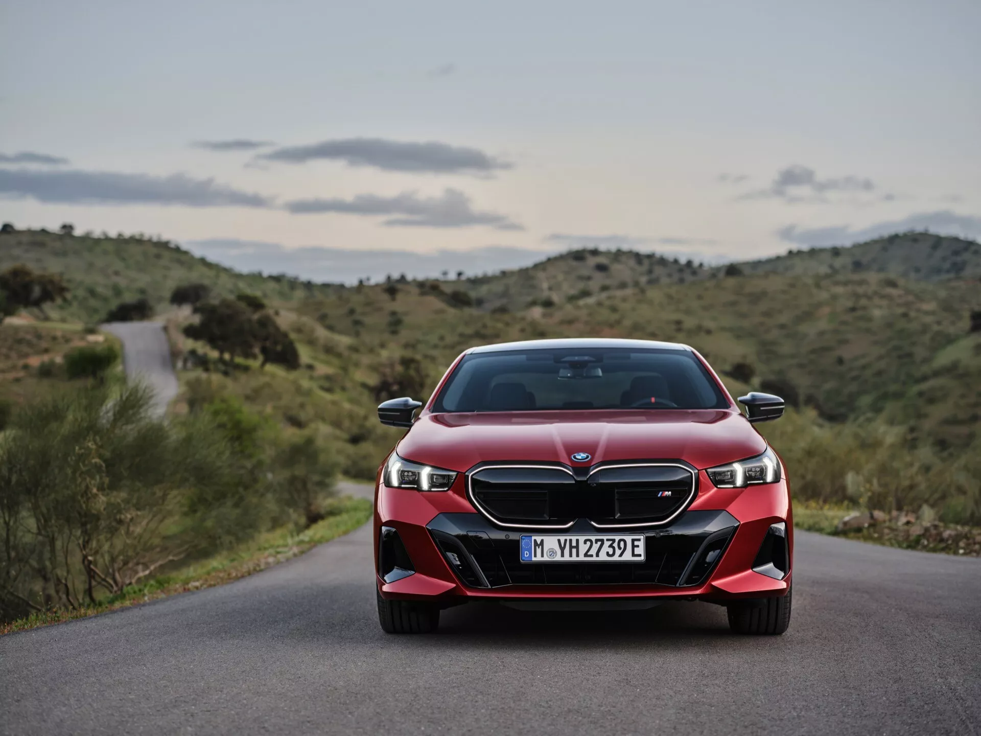 Čelný pohľad na červené BMW rady 5 (G60-G61) ročník 2023, zobrazujúci LED svetlomety, dvojitú obličkovú mriežku a športový nárazník na pozadí kľukatej cesty a kopcovitej krajiny