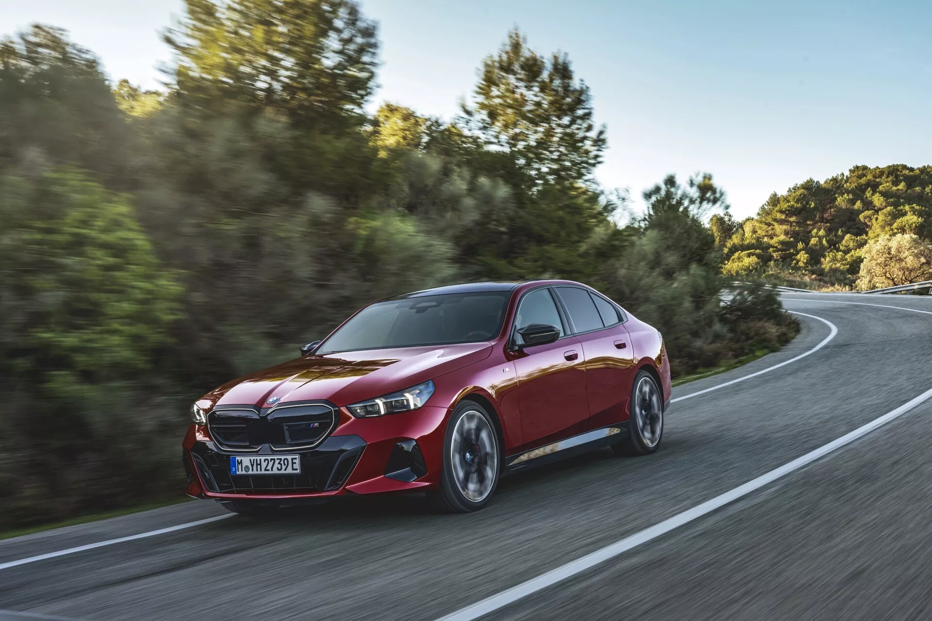 Dynamický trojštvrtečný pohľad na BMW radu 5 (G60-G61) z roku 2023 v pohybe na zakrivenej ceste uprostred vidieckej scény, zvýrazňujúci jeho metalický červený lak, charakteristickú prednú mriežku, štíhle LED svetlomety, aerodynamické bočné zrkadlá, športové kontúry, pneumatiky s nízkym profilom a viacramenné zliatinové kolesá