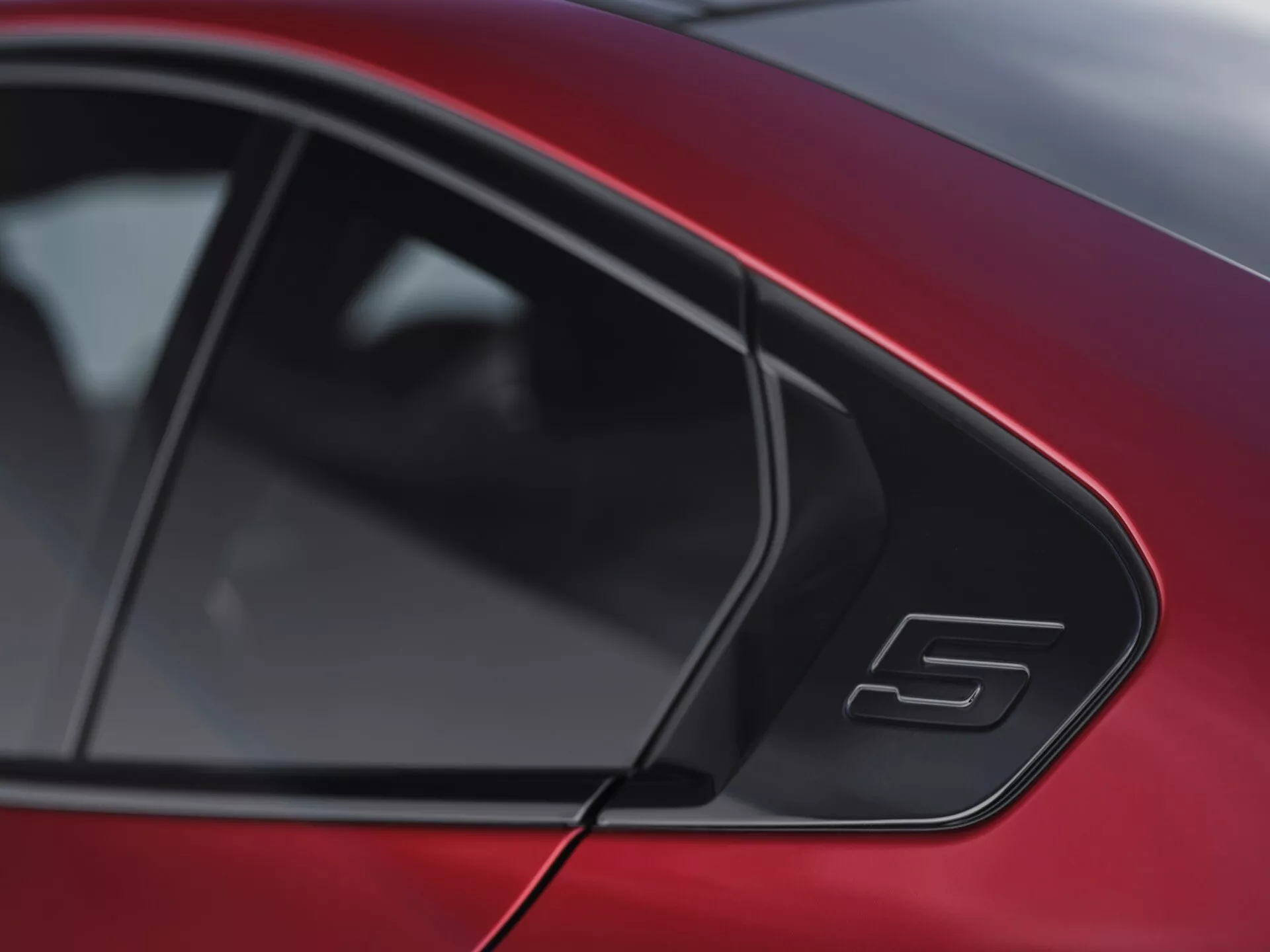 Detailný pohľad na zadné bočné okno a slúpek C červeného BMW radu 5 (G60-G61) z roku 2023 s čiernou lištou a odznakom 'S', ktorý predvádza jeho štíhlu siluetu podobnú kupé a športové detaily.