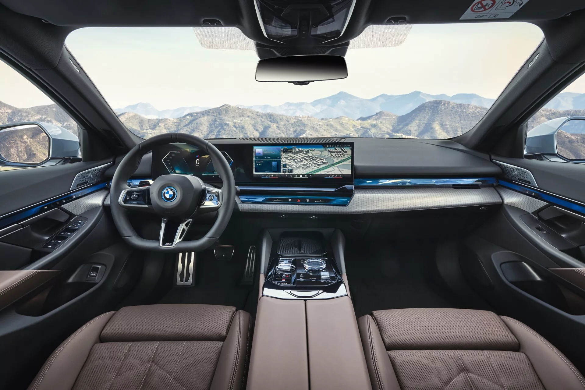Interiér BMW radu 5 (G60-G61) z roku 2023 z pohľadu zadného sedadla ukazuje prístrojovú dosku, volant, stredovú konzolu a priestor pre spolujazdca s luxusnými sedadlami vo dvojfarebnom prevedení a modrým ambientným osvetlením