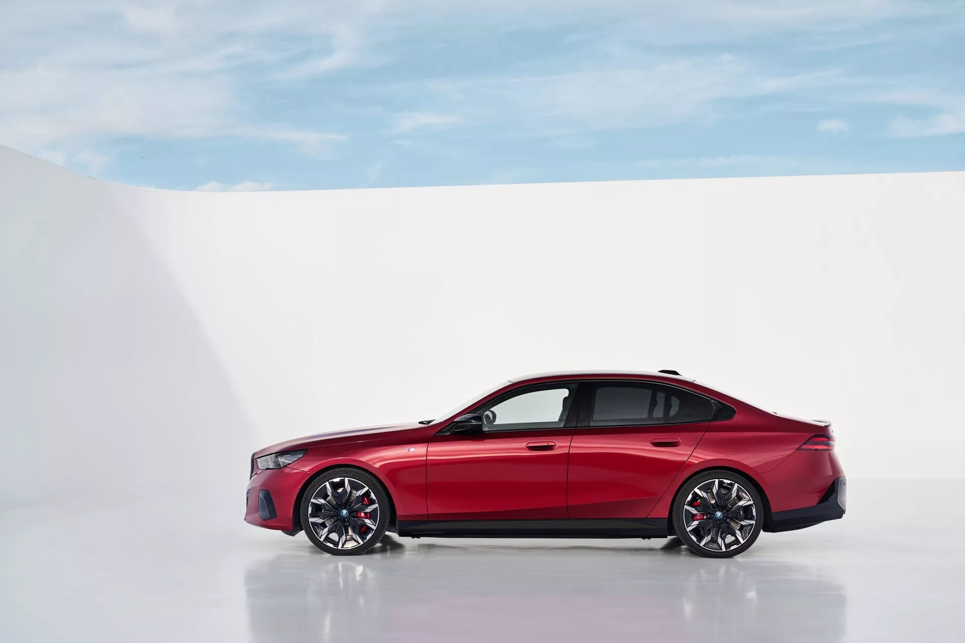 BMW radu 5 (G60-G61) rok 2023 v jasne červenej farbe, zachytené z profilu na odrazivej ploche proti bielemu pozadiu, zdôrazňujúce jeho elegantný dizajn a aerodynamické prvky, vrátane viacramenných zliatinových kolies a pneumatík s nízkym profilom.