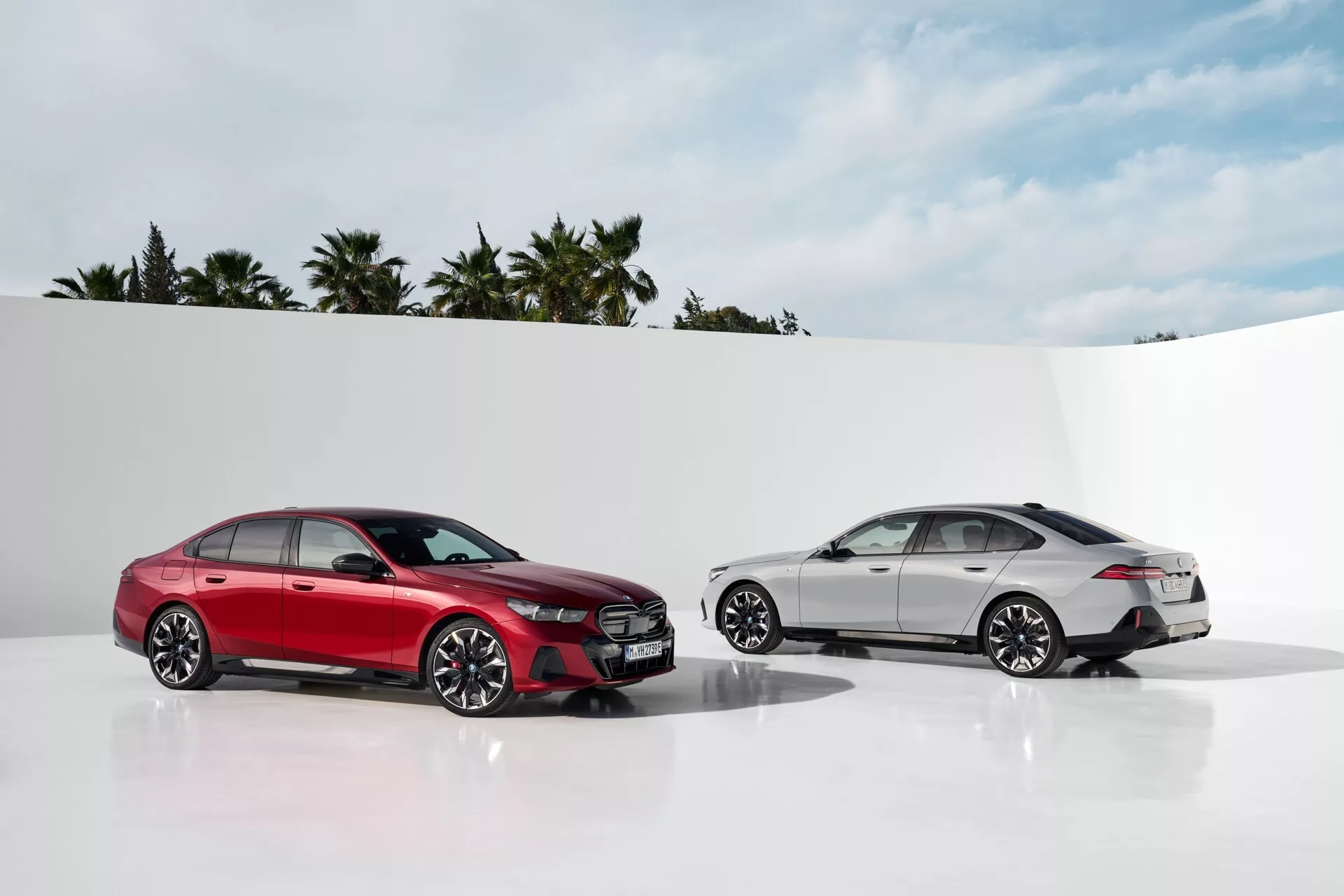 BMW radu 5 (G60-G61) 2023, červený sedan z profilu a biely sedan z trojštvrtečného zadného pohľadu na čistom bielom pozadí s palmami a jasnou oblohou