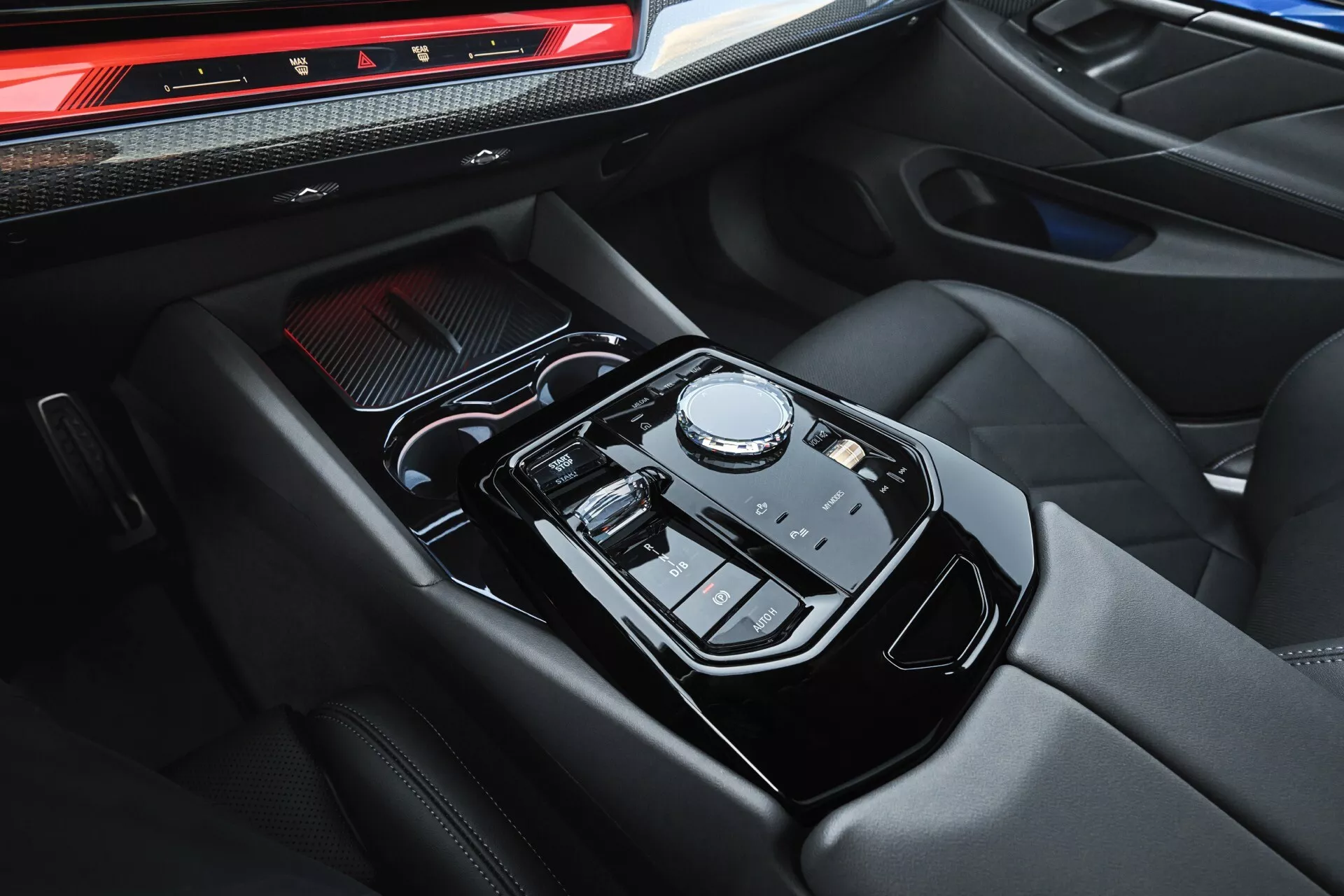 Detailný pohľad na stredovú konzolu BMW radu 5 (G60-G61) 2023 s automatickým výberom prevodov pomocou otočného voliča, dotykovým ovládaním infotainment systému, luxusnou opierkou ruky z kože s kontrastným prešívaním a ergonomickým rozložením tlačidiel a prepínačov pre režimy jazdy