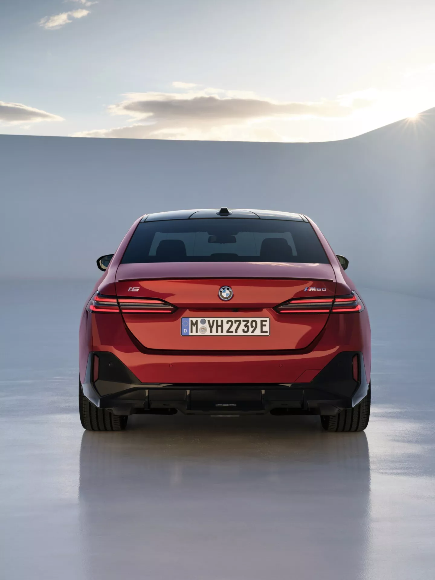 Zadný pohľad na BMW radu 5 (G60-G61) rok 2023 s červeným exteriérom, štíhlymi horizontálnymi LED zadnými svetlami a označením elektrického modelu, na pozadí s jemnou líniou obzoru a osvetlením zo štúdia alebo zamračenou oblohou