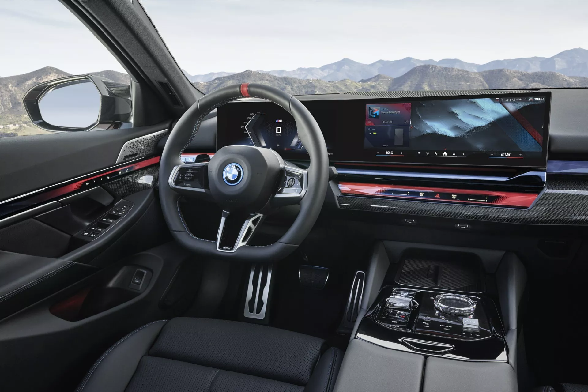 Pohľad do interiéru BMW radu 5 (G60-G61) z roku 2023 z perspektívy zadného pasažiera zobrazujúci palubnú dosku, multifunkčný volant s logom BMW, digitálny prístrojový panel, rozsiahlu obrazovku infozábavného systému, elektronickú radiaciu páku a luxusné povrchové úpravy vrátane kože a karbónových prvkov.