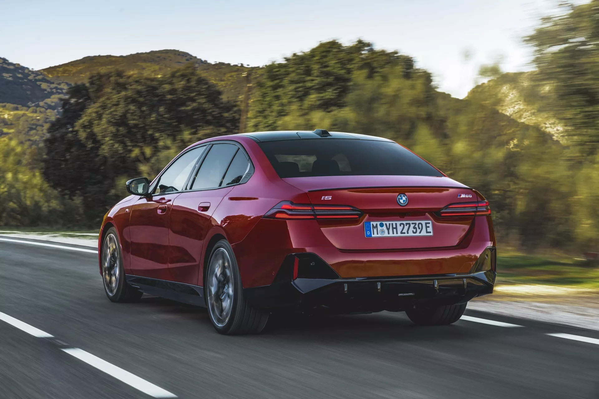 Moderný sedan BMW radu 5 (G60-G61) z roku 2023 idúci po otvorenej ceste, trojštvrtinový zadný pohľad zobrazujúci elegantné zadné svetlá, športový zadný nárazník so zabudovanými výfukovými portami a charakteristický kupé-podobný profil.