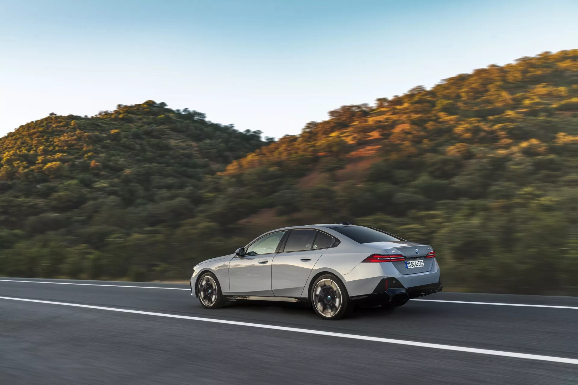 Moderný sedan BMW radu 5 (G60-G61) z roku 2023 v pohybe na zpevnenej ceste, ukazujúci svoj aerodynamický dizajn, LED zadné svetlá, zliatinové kolesá a luxusný strieborný povrch, proti pozadiu kopca s hustou vegetáciou pri západe slnka