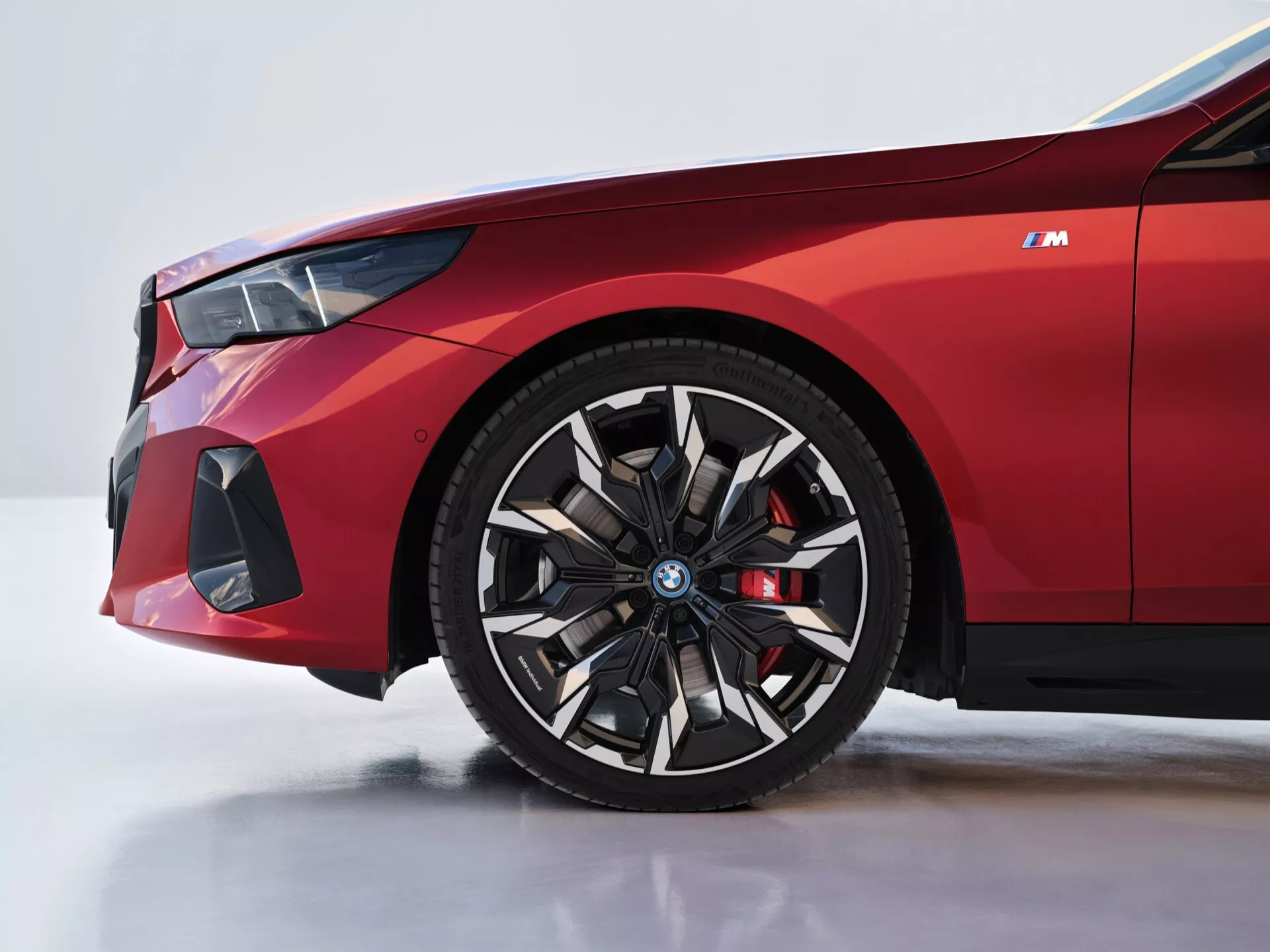 Detailný pohľad na prednú ľavú stranu BMW radu 5 (G60-G61) ročník 2023 so zameraním na koleso a blatník, s detailmi ako logo na modrom kryte stredu kolesa, červený brzdový strmienok a predný nárazník orientovaný na výkon.