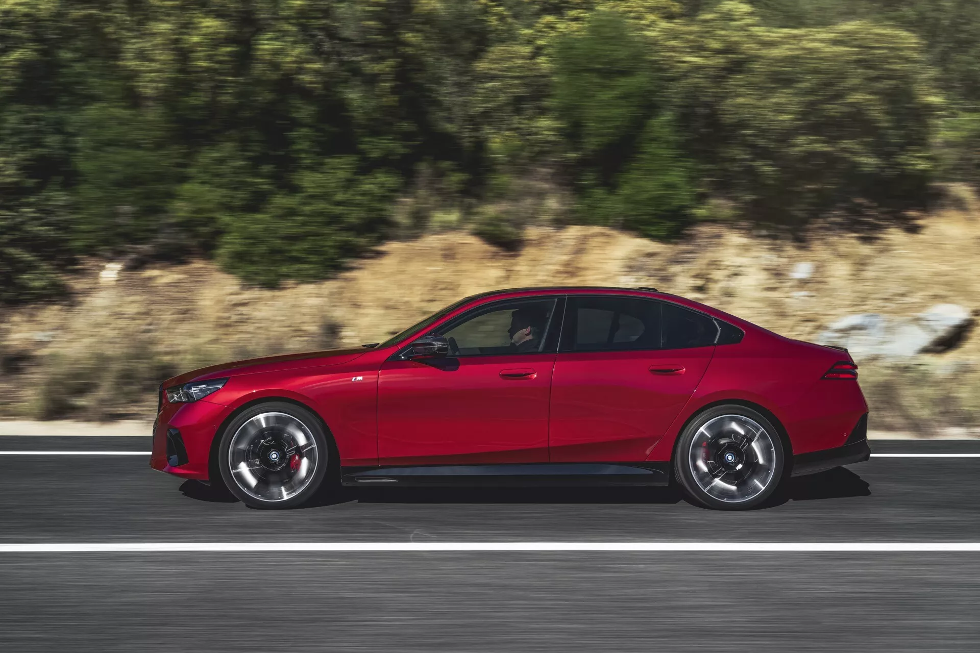 Dynamický bočný profil BMW radu 5 (G60-G61) z roku 2023 v pohybe na ceste, ktorý zdôrazňuje športový dizajn, veľké viacramenné zliatinové kolesá a lesklý červený lak zdôrazňujúci aerodynamiku a pokročilé technologické funkcie
