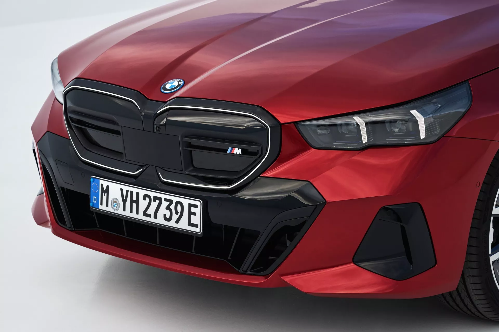Zblízka uhlový pohľad na ľavú prednú stranu červeného BMW radu 5 (G60-G61) z roku 2023 s ikonickou obličkovou mriežkou označenou ako model 'M', LED svetlometovým zoskupením a špecializovanými aerodynamickými prvkami naznačujúcimi elektrické alebo hybridné schopnosti