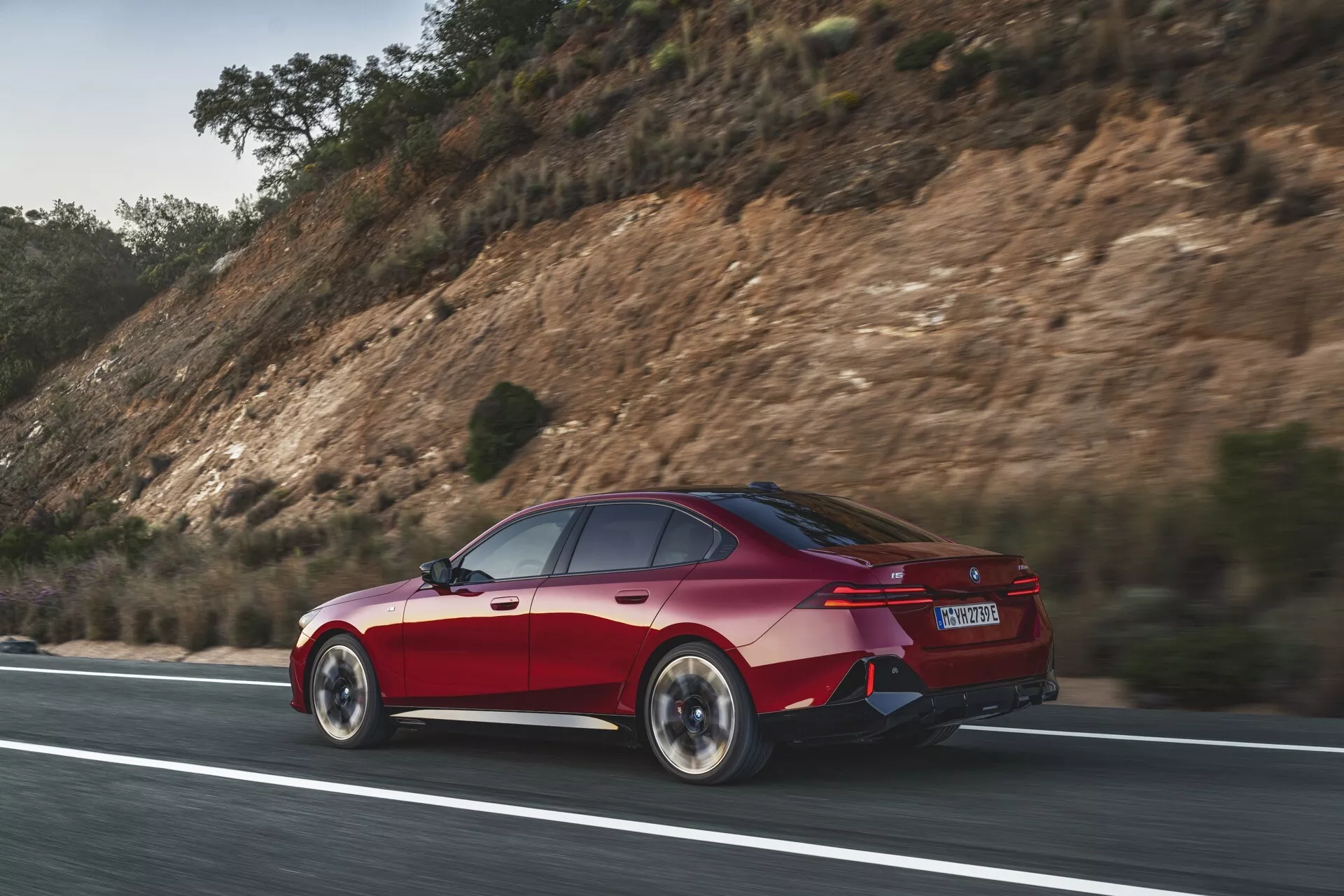 Červená štvordverová limuzína BMW radu 5 (G60-G61) v dynamickom pohybe na ceste, zachytená z bočného zadného uhla zdôrazňujúca aerodynamický dizajn auta, LED zadné svetlá, zadné koleso a nárazník