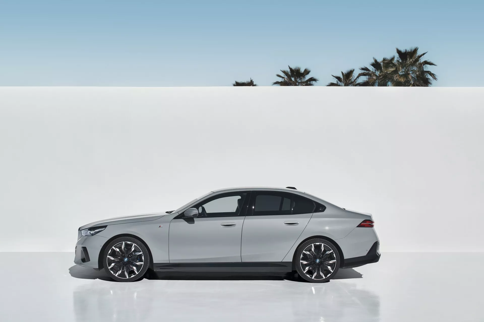 Profilový pohľad na BMW radu 5 (G60-G61) 2023 ukazuje elegantný dizajn a športovú estetiku s metalickým lakom, veľkými zliatinovými kolesami a aerodynamickou siluetou na minimalistickom pozadí s palmami