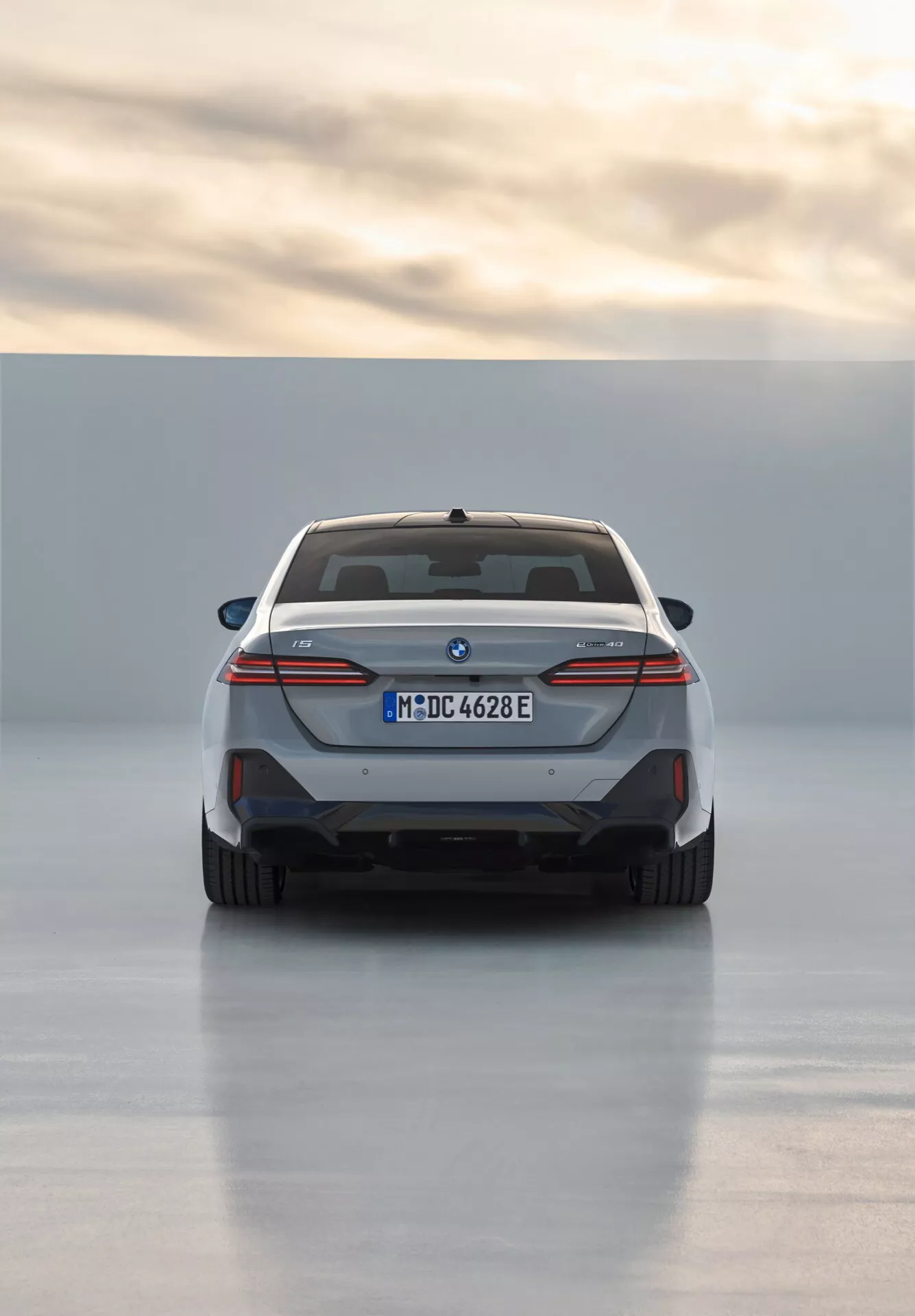 Zadný pohľad na BMW radu 5 (G60-G61) rok 2023 ukazujúci dizajn zadných svetiel na celú šírku, modré akcenty elektrického modelu, dvojité výfuky a športový zadný difúzor, registrované v Mníchove s efektom odrazeného svetla na podlahe