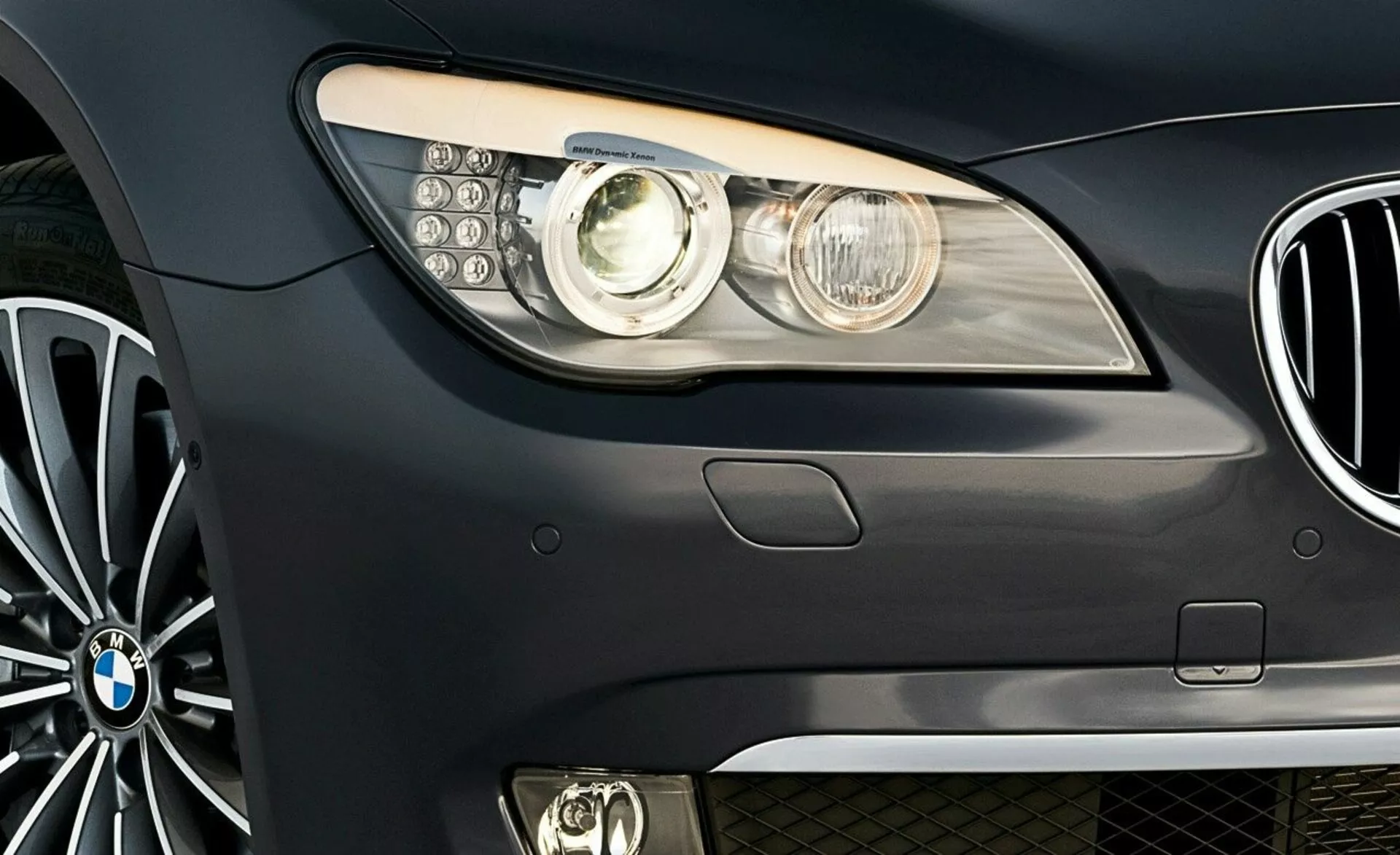 Zblízka predná bočná pohľad na BMW radu 7 z roku 2005 s emblémom značky, modernou svetlometovou jednotkou s projekčnou šošovkou a LED dennými svetlami, čiastočným výhľadom na obličkovú mriežku a viacramenným zliatinovým kolesom.