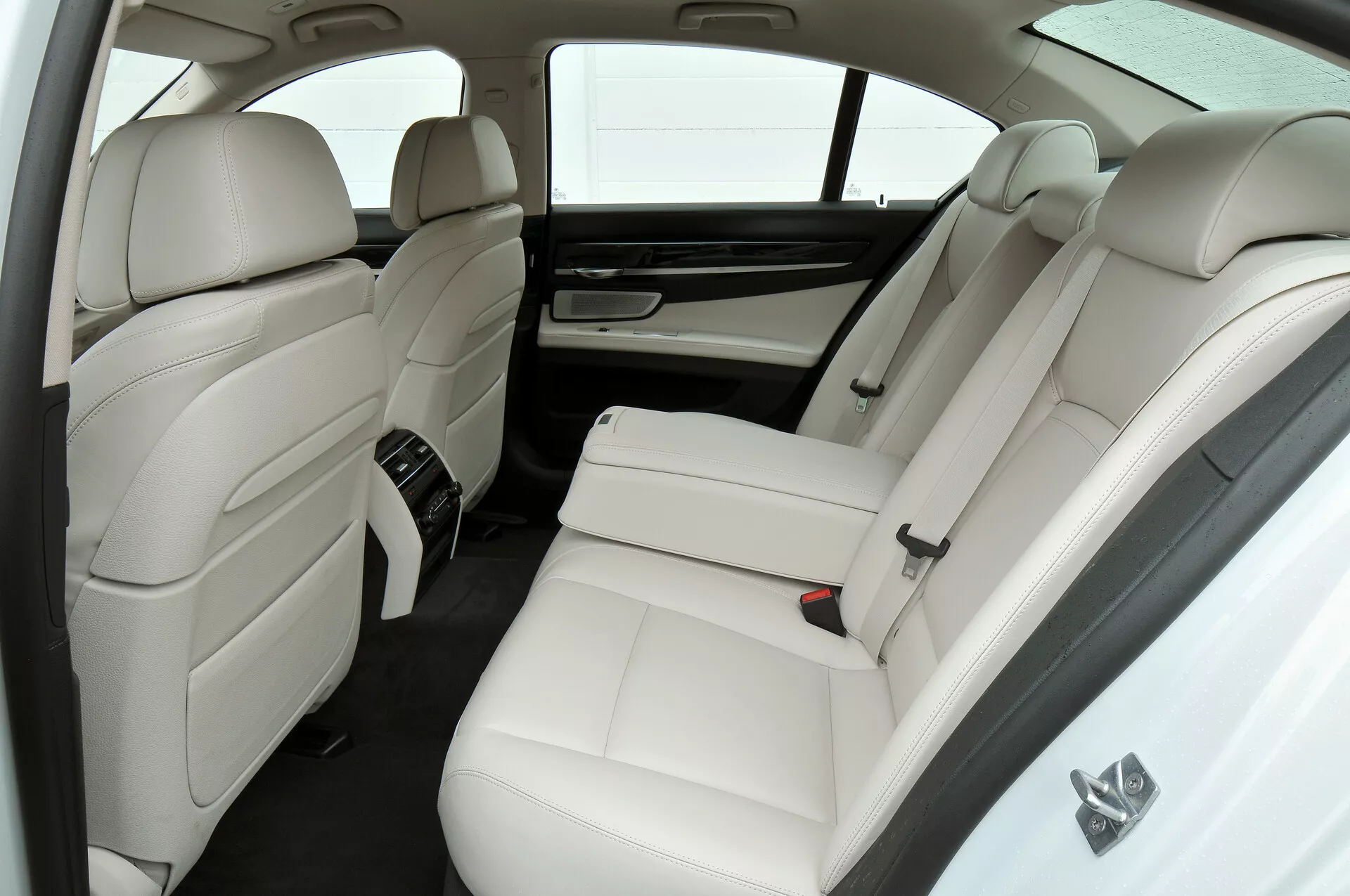 Obrázok zobrazuje luxusný interiér z bielej kože vo voze BMW radu 7 (facelift 2012, F01-F02), so zameraním na zadný pasažiersky priestor z mierne zvýšenej perspektívy. Sú tu dve samostatné zadné sedadlá s opierkami hlavy a bočnými oporami, stredová konzola s ovládacími prvkami, zadné vetracie otvory klimatizácie, slnečná clona na zadnom okne, integrované opierky hlavy na operadlách predných sedadiel a minimalistická farebná paleta v bielej a tmavých tónoch.