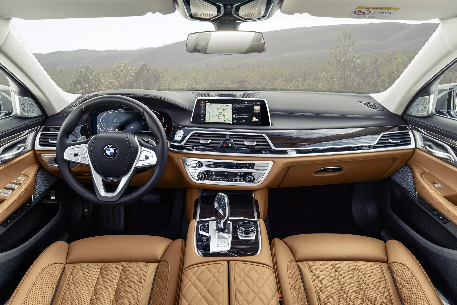 Obrázok ukazuje interiér BMW radu 7 z roku 2019 (facelift G11/G12) z pohľadu zadných sedadiel, zdôrazňujúc detailný a luxusný dizajn prednej kabíny. Moderný volant s multifunkčnými tlačidlami a výrazným emblémom BMW dopĺňa elegantná prístrojová doska s centrálnym dotykovým infotainment displejom. Stredová konzola obsahuje automatickú radiacu páku, touchpad a rôzne dynamické ovládacie systémy, doplnené svetlými koženými sedadlami s prešívaním, čo naznačuje prémiovú úroveň výbavy. Dverové panely odrážajú tento luxus s koženým obložením a drevom, poskytujúc ambientné osvetlenie, čo naznačuje, že pokročilé technologické a komfortné prvky sú integrované v celom vozidle, vrátane strešného okna pre ďalšie prirodzené svetlo.