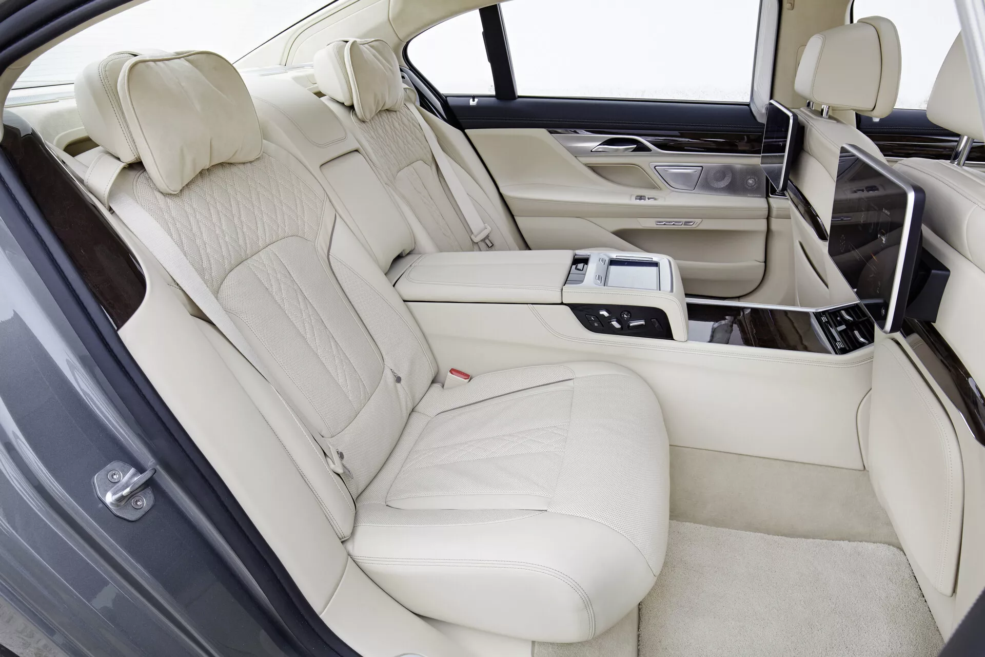 Interiér BMW radu 7 (G11-G12) model 2015, zobrazujúci luxusný priestor zadných sedadiel s ľahkým krémovým koženým čalúnením, tvarované zadné sedadlá pre dvoch pasažierov oddelené stredovou konzolou. Konzola zahrnuje technologické vybavenie, ako je ovládací panel, klimatizácia, vyhrievanie alebo chladenie sedadiel, mediálne možnosti a držiaky na poháre. Zadné zábavné obrazovky sú integrované do zadnej časti predných sedadiel, s drevenými obloženiami na konzole a dverách, za jasných svetelných podmienok.