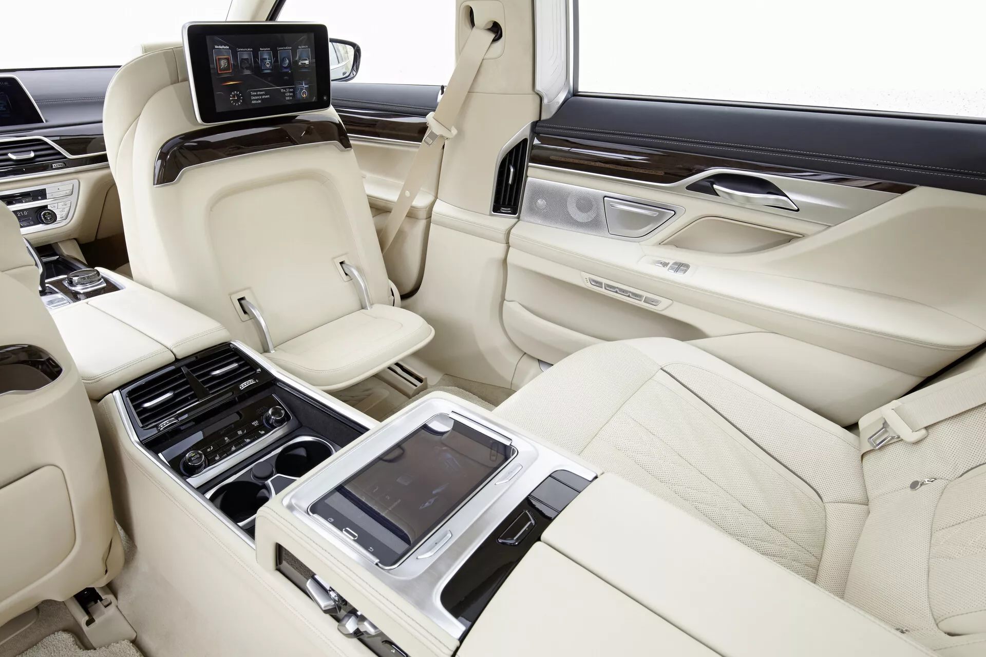 Obraz interiéru zadnej kabíny BMW 7er-reihe (G11-G12) z roku 2015, zachytávajúci priestrannú a luxusnú sedaciu plochu z pravostranného pohľadu. Svetlé kožené sedadlá v usporiadaní 2+1, s tvarovaným dizajnom pre pohodlie a podporu, opierkami hlavy a individuálnymi schopnosťami nakláňania sú viditeľné. Centrálna konzola s rôznymi ovládacími prvkami a prípadne dotykovým rozhraním pre nastavenie sedadiel, klimatizáciu a multimédiá je zdôraznená. Dverné panely obsahujú drevené obloženie a kartáčované kovové akcenty s ďalšími ovládacími prvkami. Na zadnej strane predných sedadiel sú namontované jednotlivé monitory pre zábavu a za centrálnou konzolou je umiestnený ovládací panel s displejom. Interiér vozidla ukazuje pokročilé nastavenie sedadiel, klimatizáciu a zábavné systémy v prvotriednych kožených, drevených a kovových úpravách.