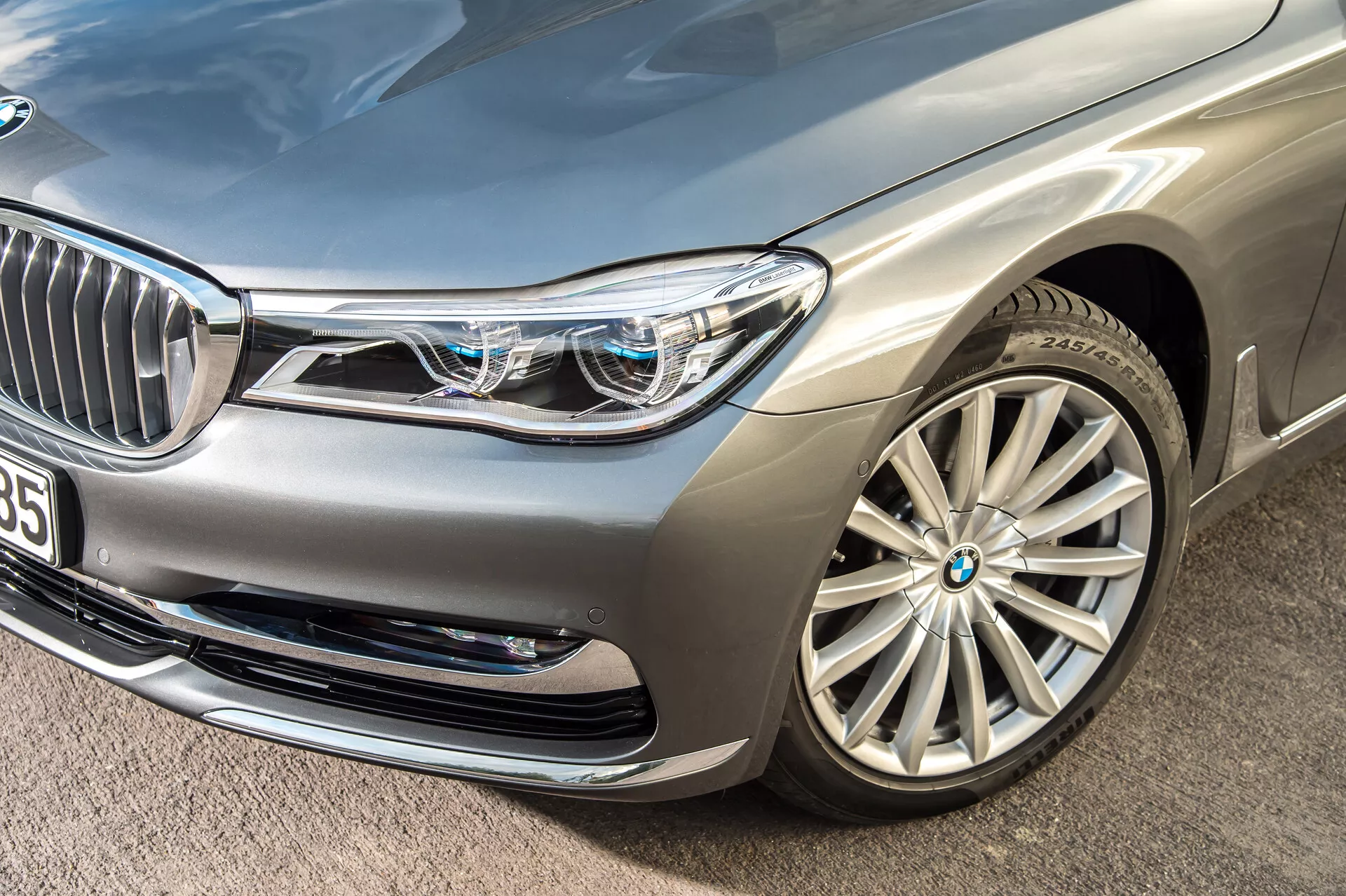 Detailný šikmý záber na strieborný luxusný sedan BMW 7er-reihe 2015 (G11-G12), zameraný na prednú ľavú časť vozidla. Zvýrazňuje predný nárazník, ľavý predný svetlomet, kapotu, blatník a viaclúčové zliatinové koleso. Zložité LED svetlomety s charakteristickým denným svietením a malými modrými štvorčekmi naznačujúcimi pokročilé osvetľovacie systémy. Čiastočne viditeľná predná mriežka s chrómovanými vertikálnymi lamelami a blízka kamera alebo senzor naznačujú technológiu asistenta vodiča. Nasávacie otvory na prednom nárazníku a metalický lak ďalej zdôrazňujú luxusný a aerodynamický dizajn vozidla.
