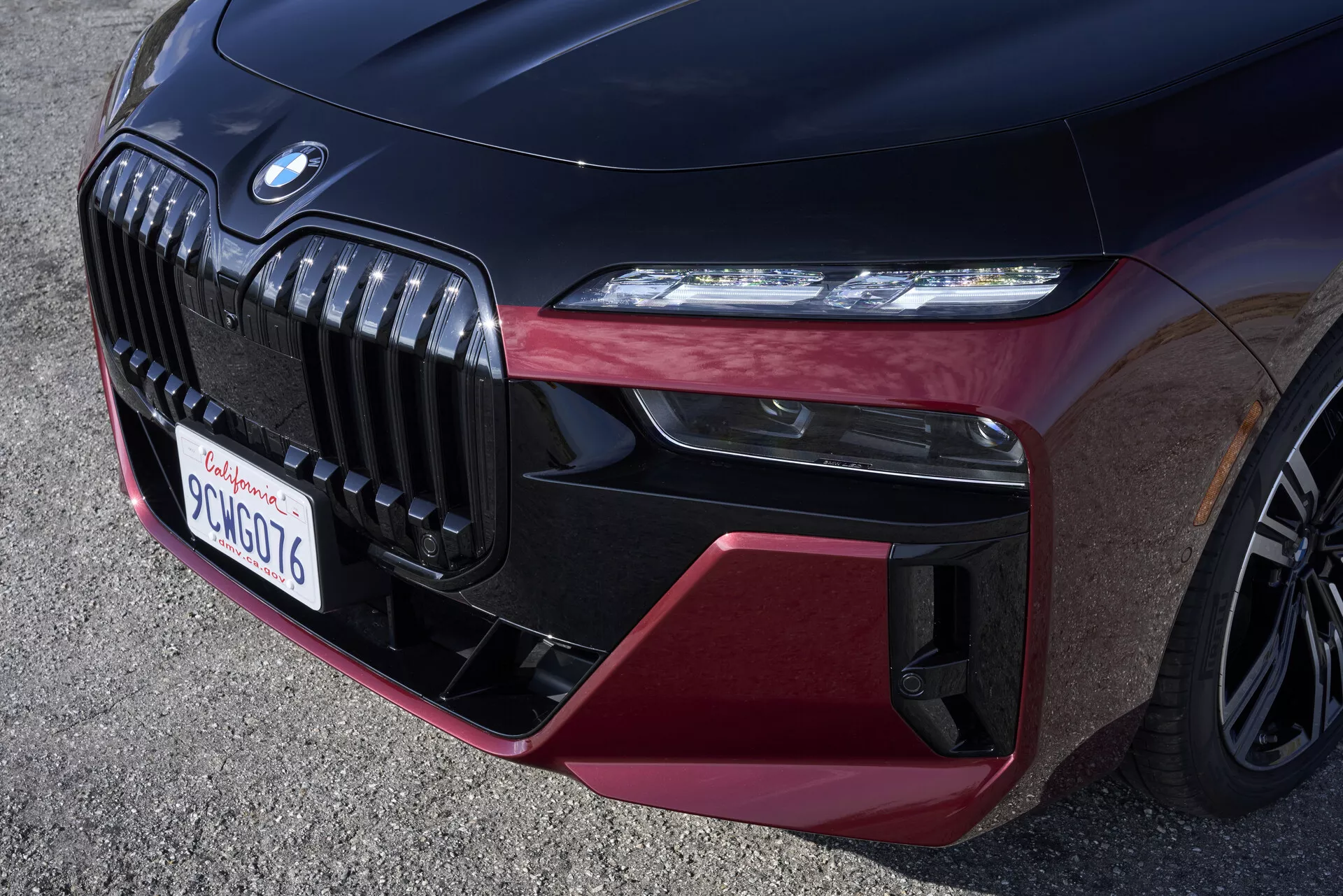 Detailný pohľad na prednú časť BMW 7er rady 2023 (G70), ktorý zvýrazňuje chrómovanú dvojitú obličkovú mriežku, elegantný LED svetlomet a dynamický predný nárazník s lesklými čiernymi a bordovými prvkami, zdôrazňujúc luxus a športový charakter. Nízký uhol odhaľuje viacramenné zliatinové koleso v rohu, ukazujúc technicky vyspelé a výkonné prvky vozidla.