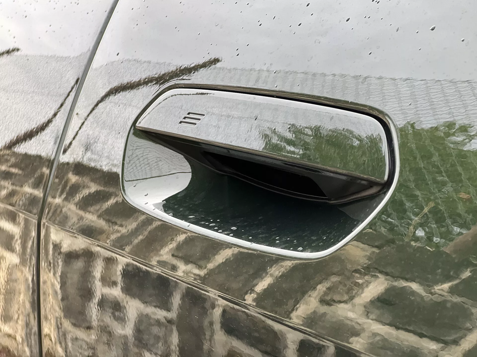 Detailný pohľad na bokové zrkadlo na strane vodiča BMW i4 2021 ukazuje jeho aerodynamický dizajn, čierne puzdro s jemnou horizontálnou textúrou, strieborný lem, povrch odolný voči poveternostným vplyvom s kvapkami vody a jeho sklápaciu funkciu odrážanú na sklá spekanej dažďom.