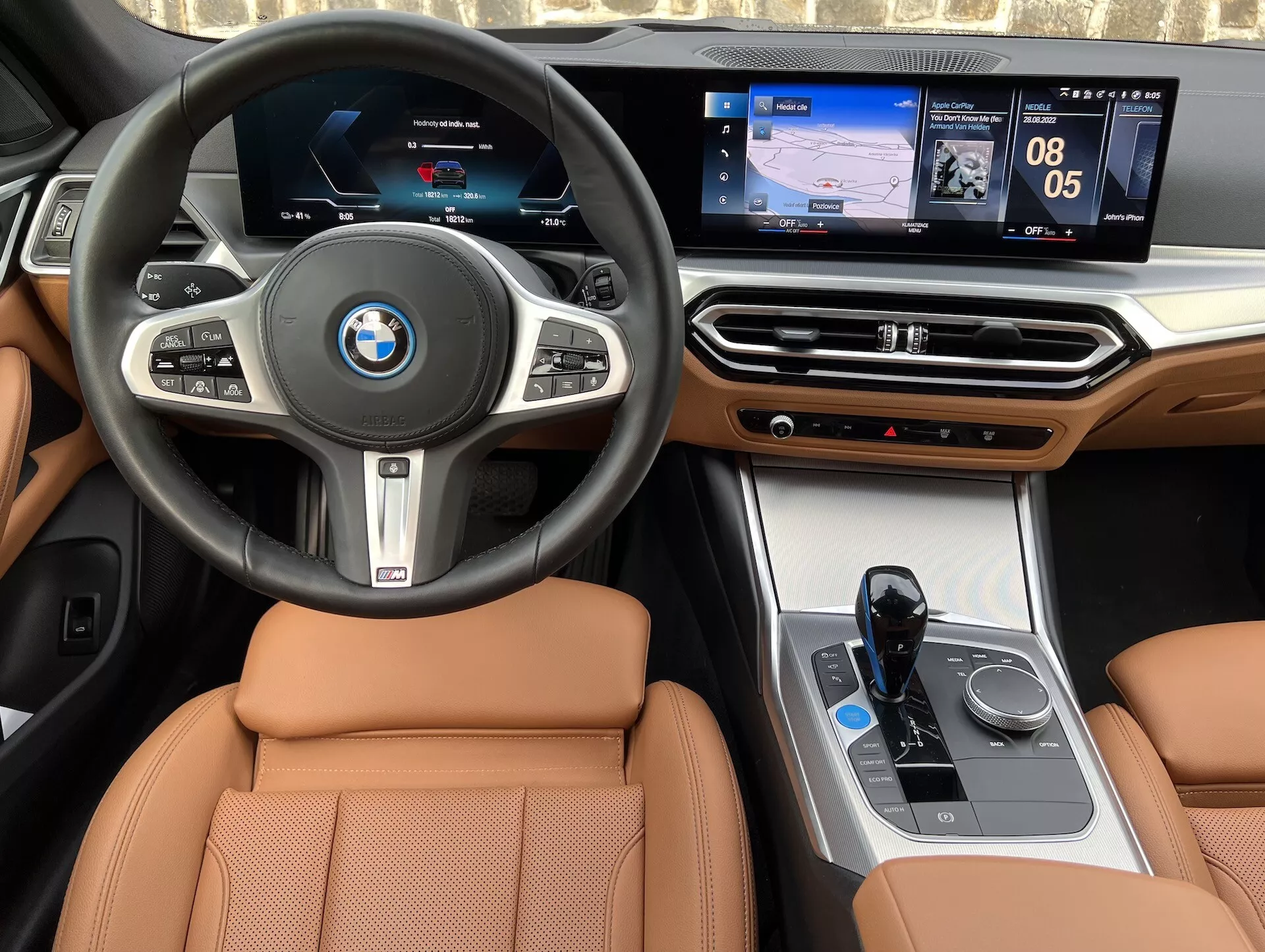 Interiérový pohľad na BMW i4 2021, obsahujúci centrálny volant s logom BMW, multifunkčné tlačidlá, digitálny prístrojový panel, dotykový centrálny informačný systém zobrazujúci navigáciu a médiá, luxusné drevené akcenty na palubnej doske, rozhranie klimatizácie, elektronickú radicu páku, ovládač iDrive a sedadlá potiahnuté svetlou kožou.
